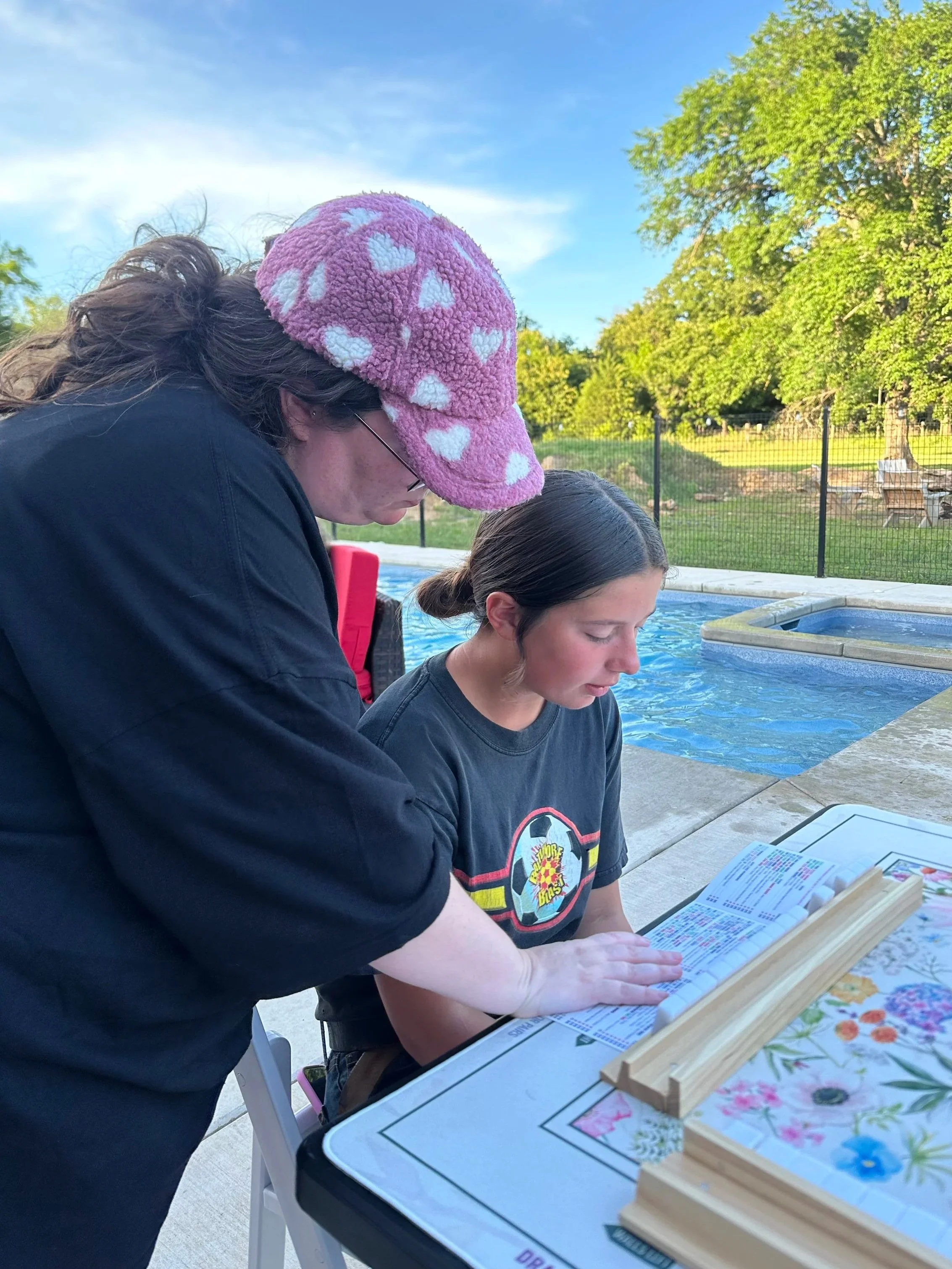 A woman with dark hair in a bun and wearing a black shirt is looking at a menu on a table next to a pool. An older woman with glasses and a pink fuzzy hat with white hearts is standing beside her, leaning over and looking at the menu. The table has a colorful floral tablecloth and a wooden puzzle. In the background, there are green trees, a fence, and a clear blue sky.