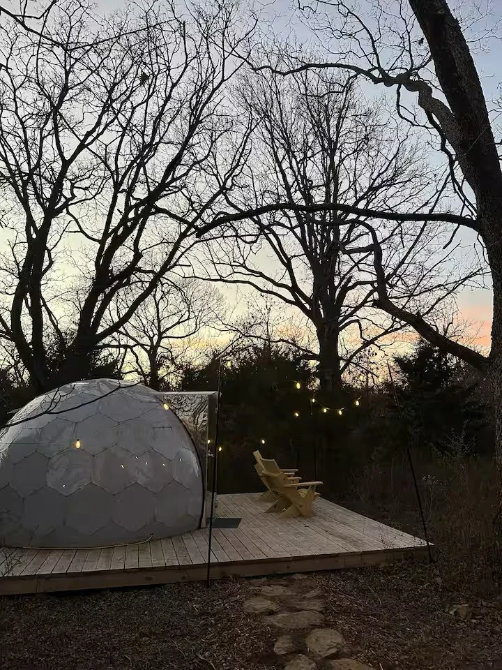 A cozy outdoor setting on a wooden deck with two wooden chairs, string lights hanging in the trees, a tent, and leafless trees in the background during dusk.