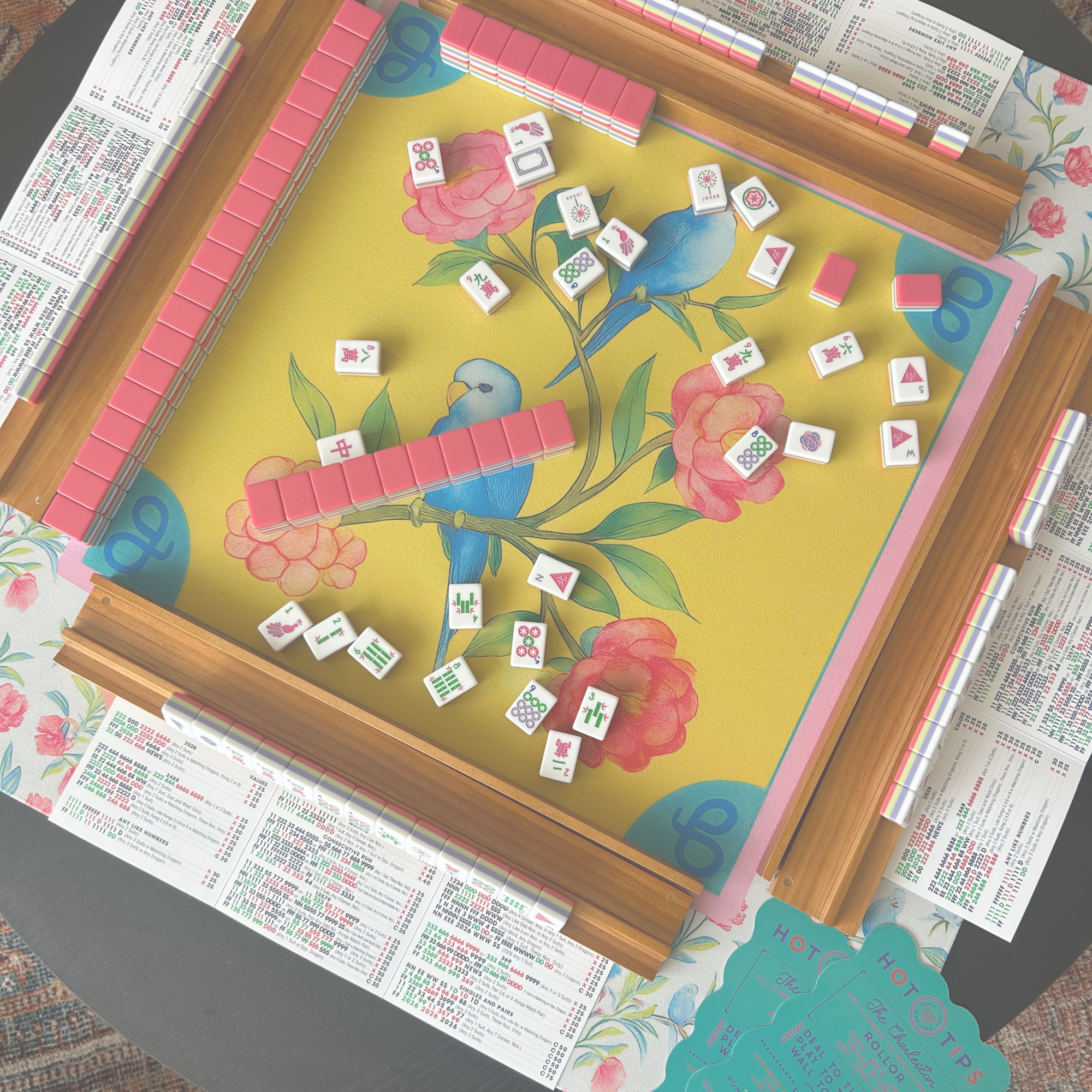 A completed Rummikub game with tiles on a floral tablecloth, surrounded by lottery or bingo tickets and pamphlets.