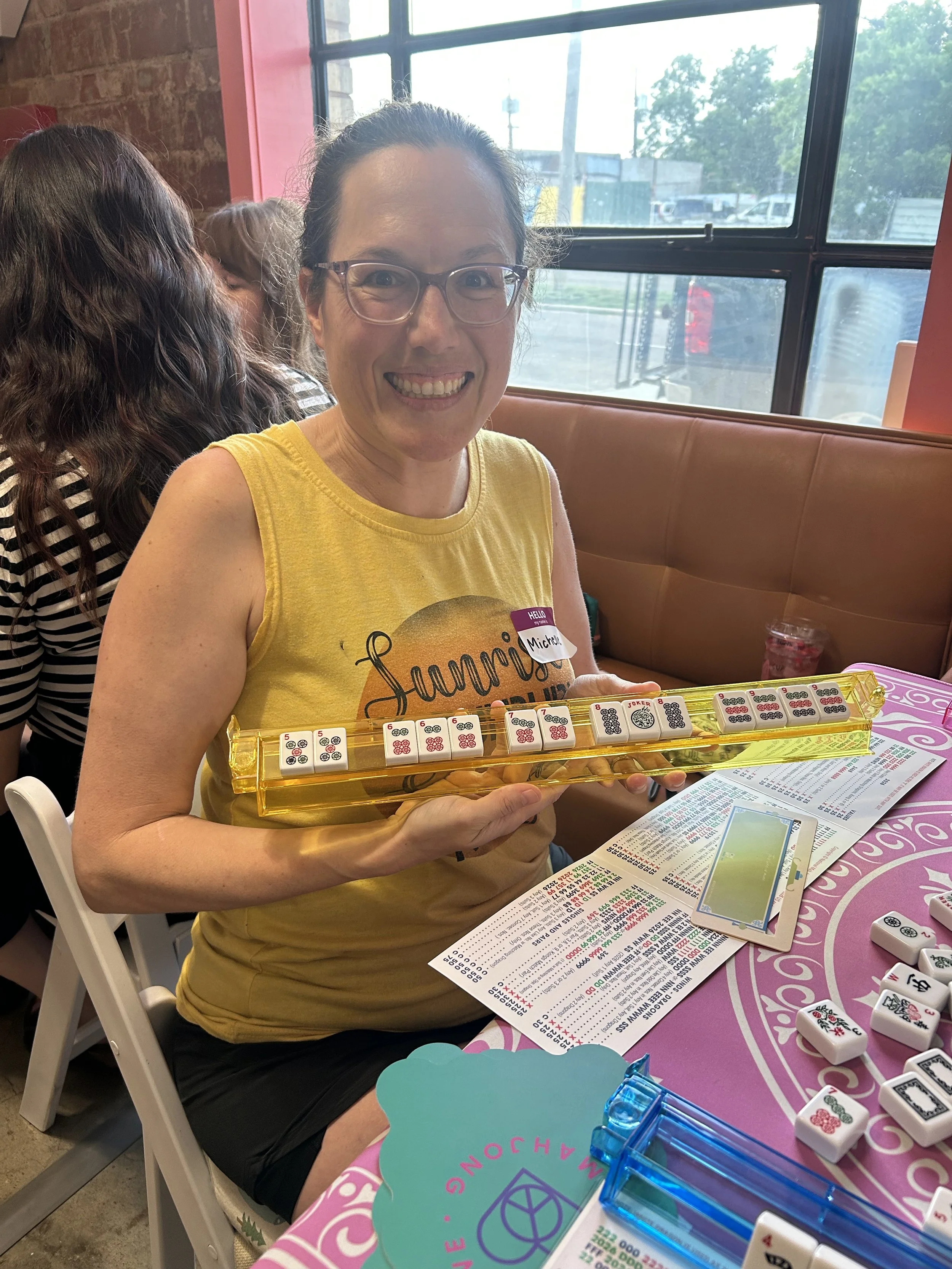 A woman smiling at the camera while holding a Mahjong tile game in a brightly lit restaurant or café. She wears glasses, a yellow sleeveless shirt, and has a name tag that says 'Michelle.' There are other people in the background, and the table has v
