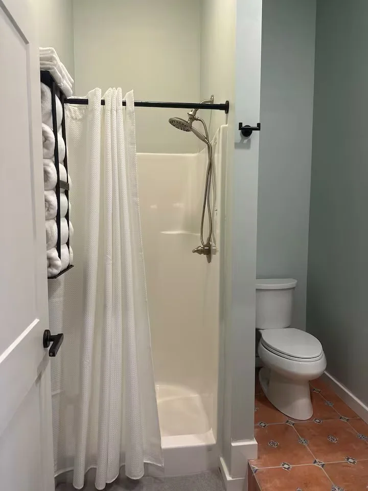 A small bathroom with a shower stall, a curtain, a toilet, and a wall-mounted shelf with rolled-up towels.