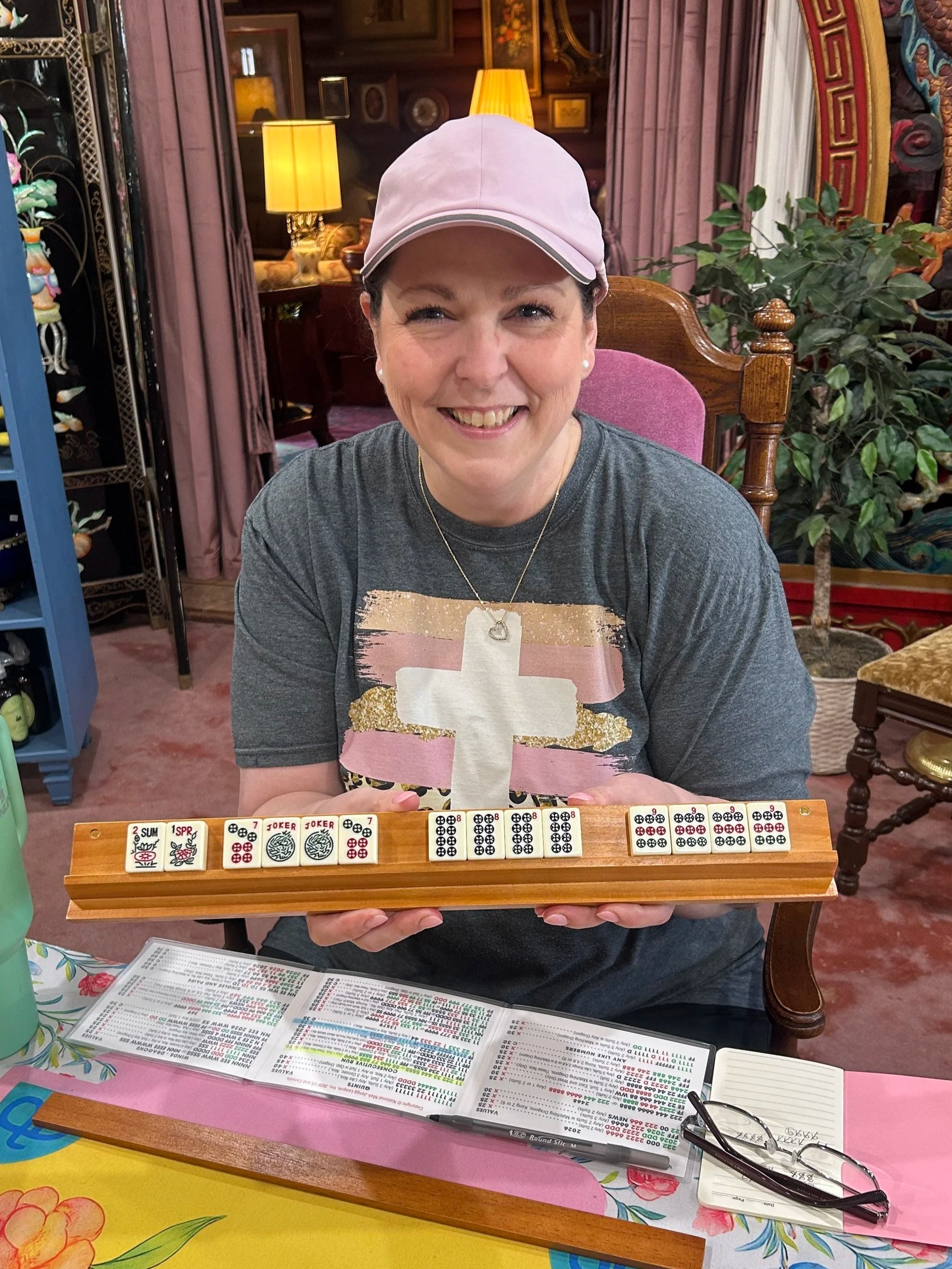 A woman smiling and sitting at a table with mahjong tiles in front of her, holding a mahjong tile. The table has various papers, a pair of glasses, and a small notepad.