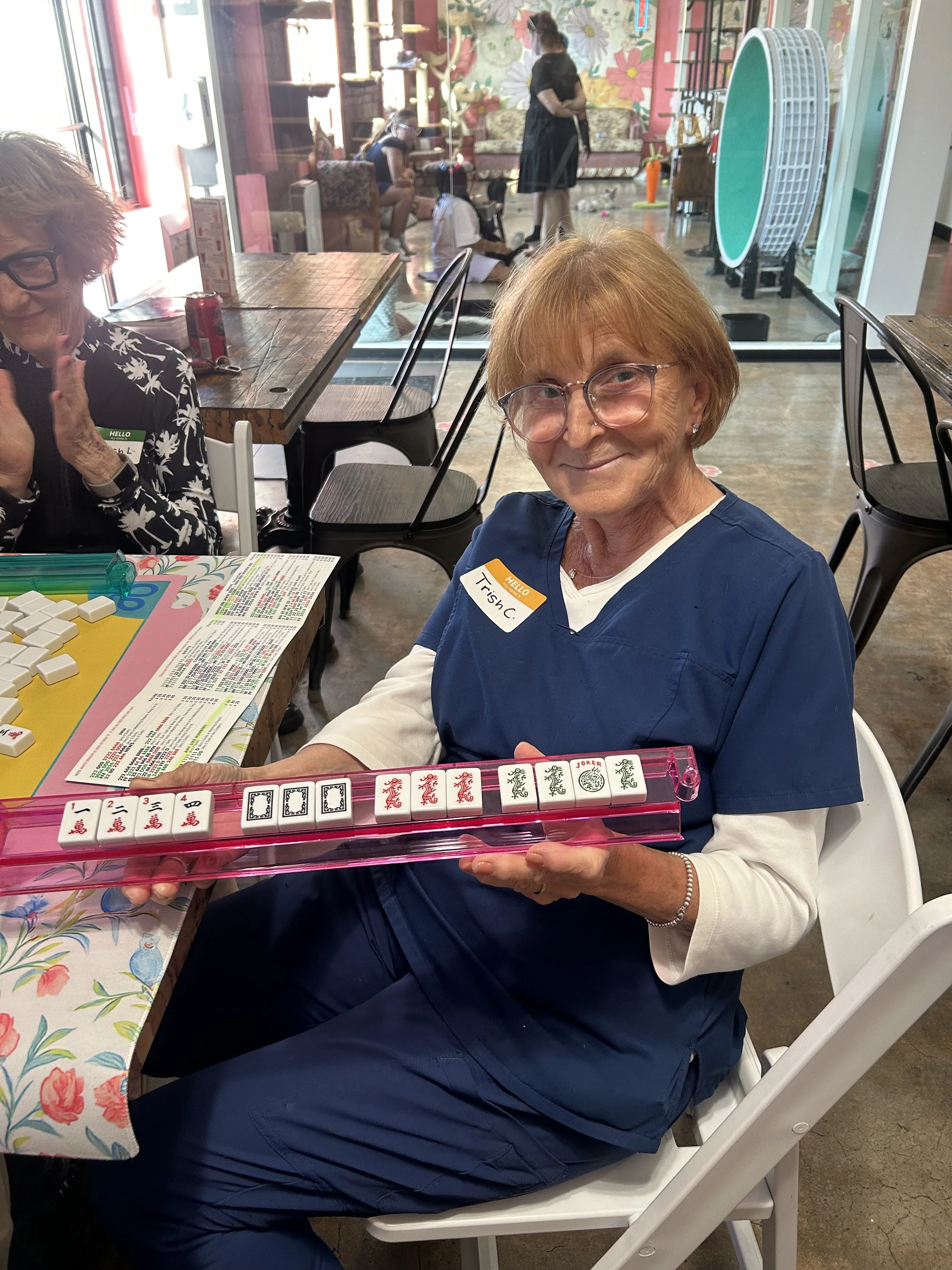 An elderly woman wearing glasses and a navy medical uniform, with a name tag that says Trish C., smiling and holding a mahjong tile rack with white tiles featuring red, black, and green designs. She is seated at a table with a colorful tablecloth.