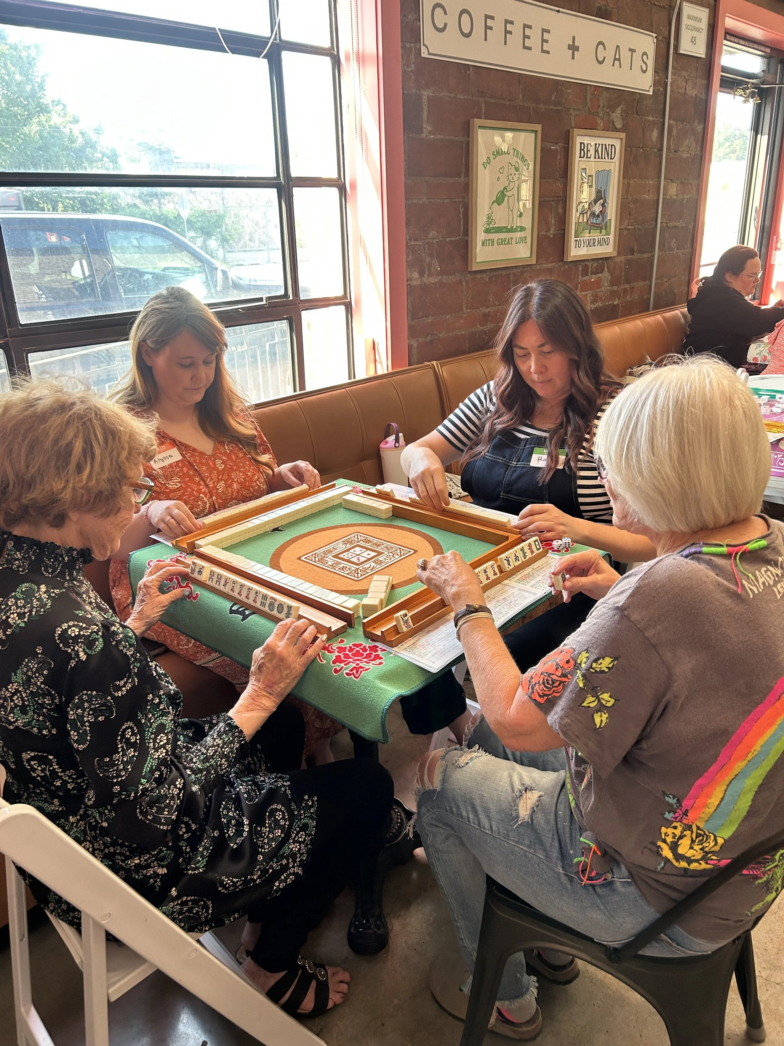 Four women playing mahjong at a table inside a cafe with a sign that reads 'Coffee + Cats' on a brick wall in the background.