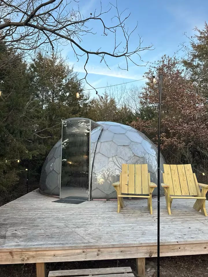 A small outdoor wooden deck with two yellow wooden chairs, a transparent geodesic dome structure, and trees in the background under a blue sky.