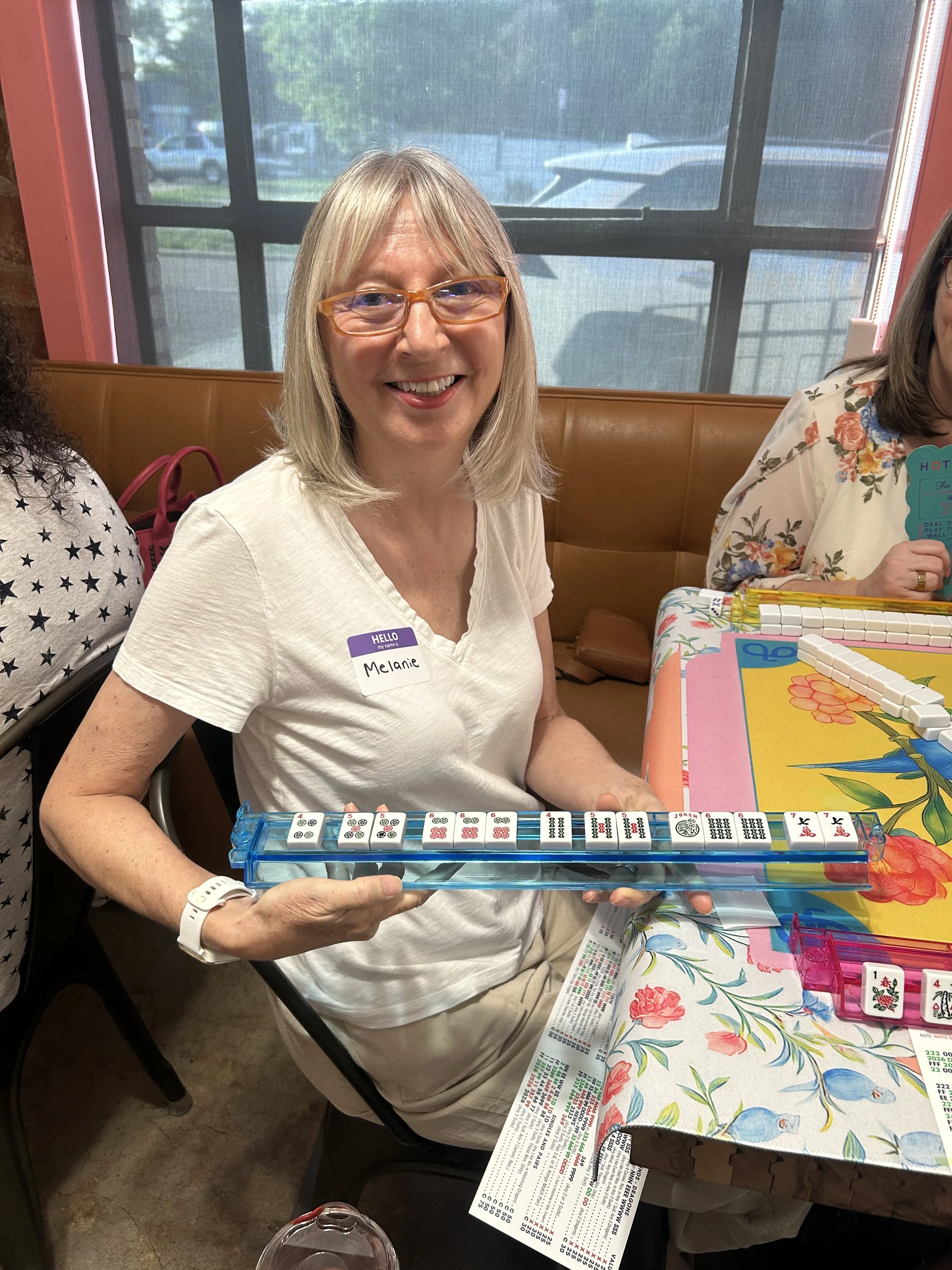 A woman with glasses and a name tag that says "Melanie" is sitting at a table with a colorful tablecloth, playing a game with domino tiles. She is smiling and holding a domino set, with other domino tiles and a score sheet on the table.