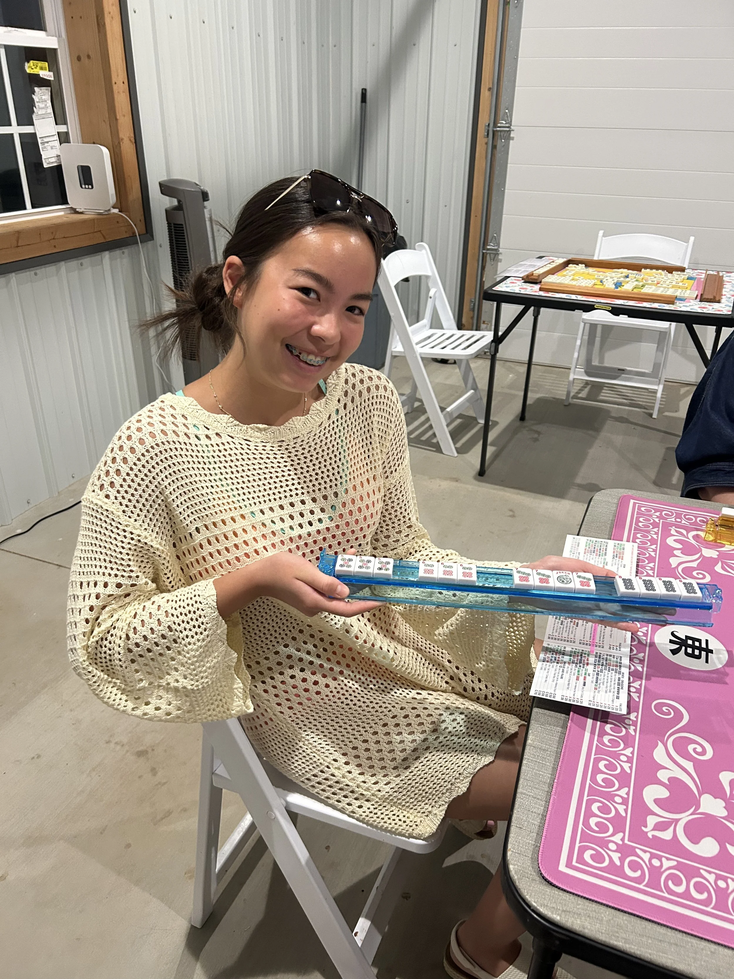 A young woman with sunglasses on her head, wearing a light-colored crochet top, sitting at a table and holding a Mahjong game set. She is smiling at the camera. The background shows a garage with a metal wall, a window, some chairs, and other Mahjong