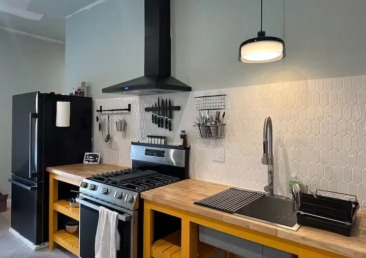 Modern kitchen with black refrigerator, stainless steel stove, wooden countertops, yellow open shelves, white textured tile backsplash, hanging light, black range hood, and kitchen utensils.