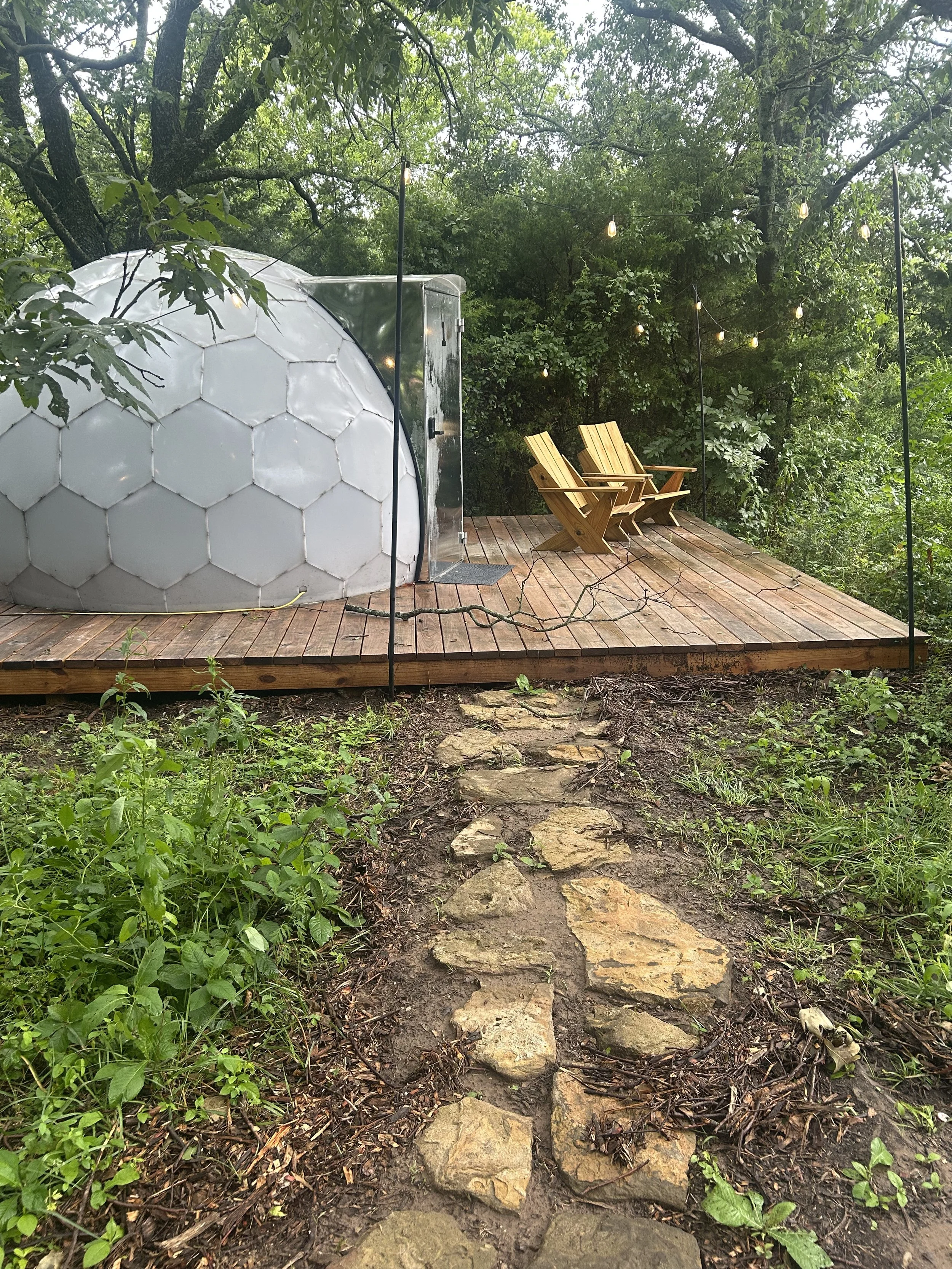 A wooden deck with two Adirondack chairs in a forested area, string lights hanging above, and a small dome-shaped structure on the left side.
