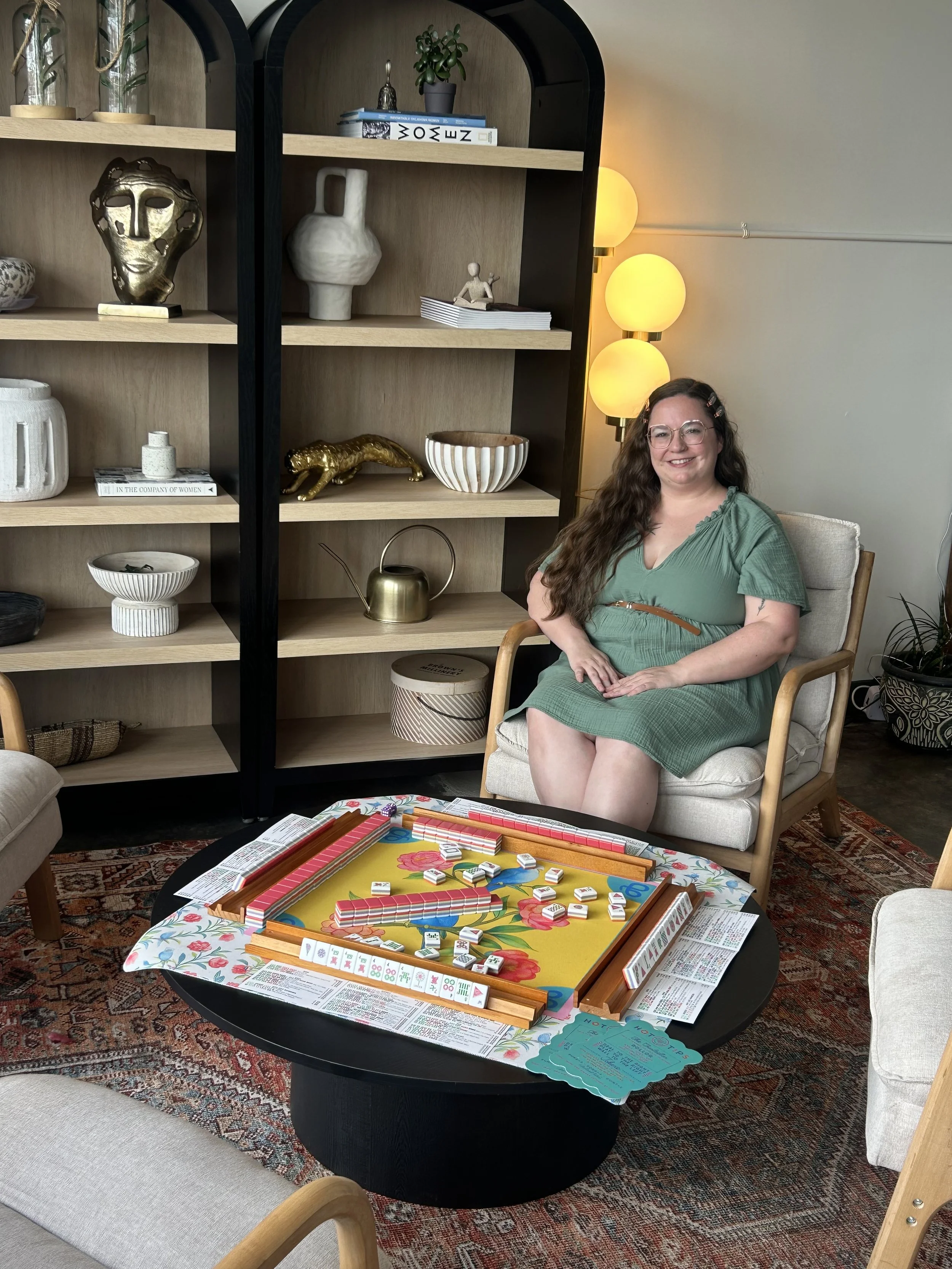 A woman with long brown hair, glasses, and a green dress sits in a chair smiling at a table with a colorful board game set up and instruction sheets. Behind her is a shelf with decorative items and a lamp with three round yellow bulbs.