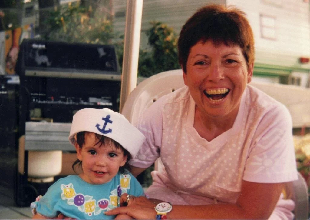 A woman and a young girl sitting closely together outdoors. The girl is wearing a sailor hat with an anchor emblem and a colorful dress. The woman is smiling broadly, wearing a light pink shirt with white polka dots. There is a grill and greenery in the background.