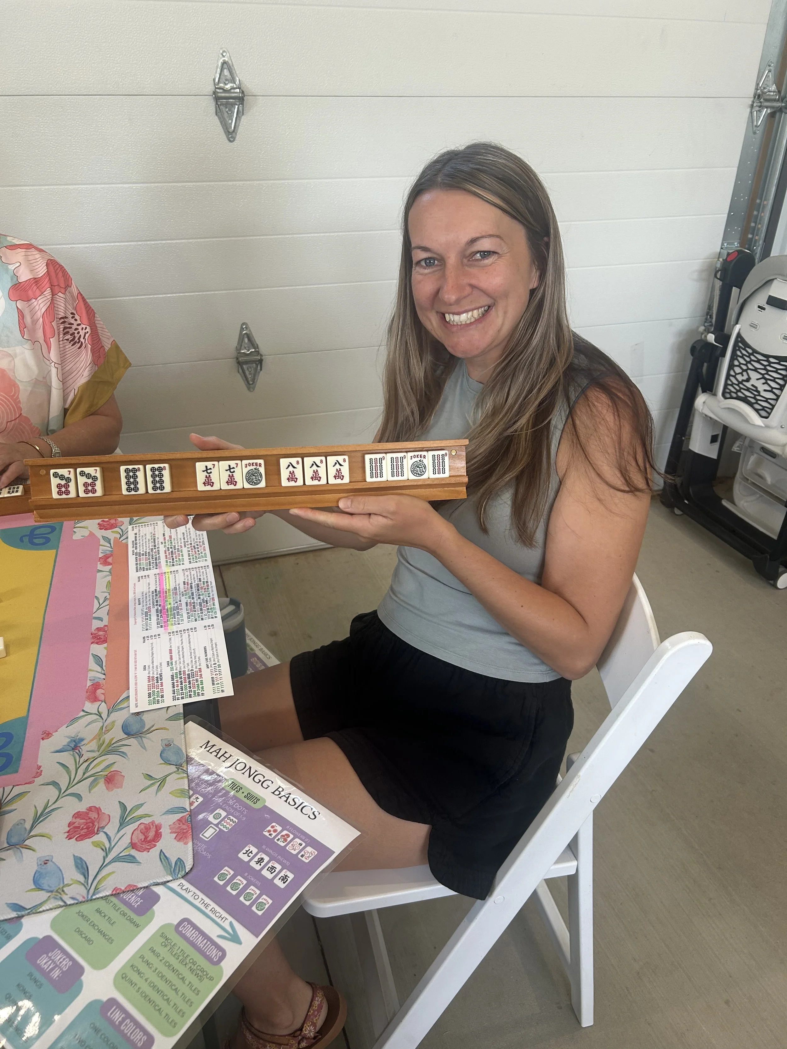 A smiling woman with long brown hair holding a mahjong set in a garage with a white door in the background. She is sitting on a white chair, wearing a sleeveless gray top and black shorts. There are colorful papers and mahjong instructions on the tab