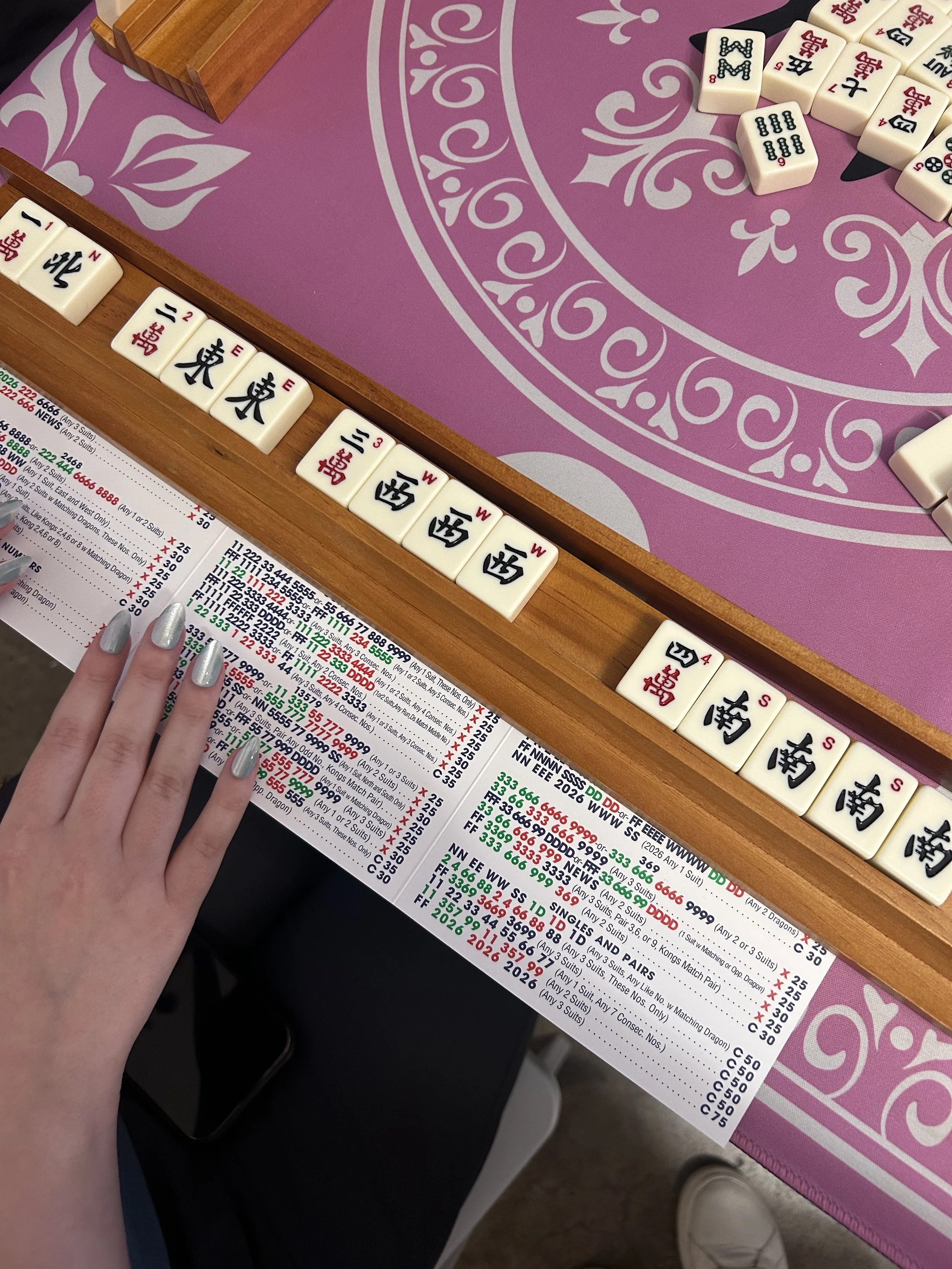 Close-up of Mahjong tiles on a pink tablecloth next to a betting slip with numbers and text. A person's hand with silver nail polish is visible near the bottom left of the image.