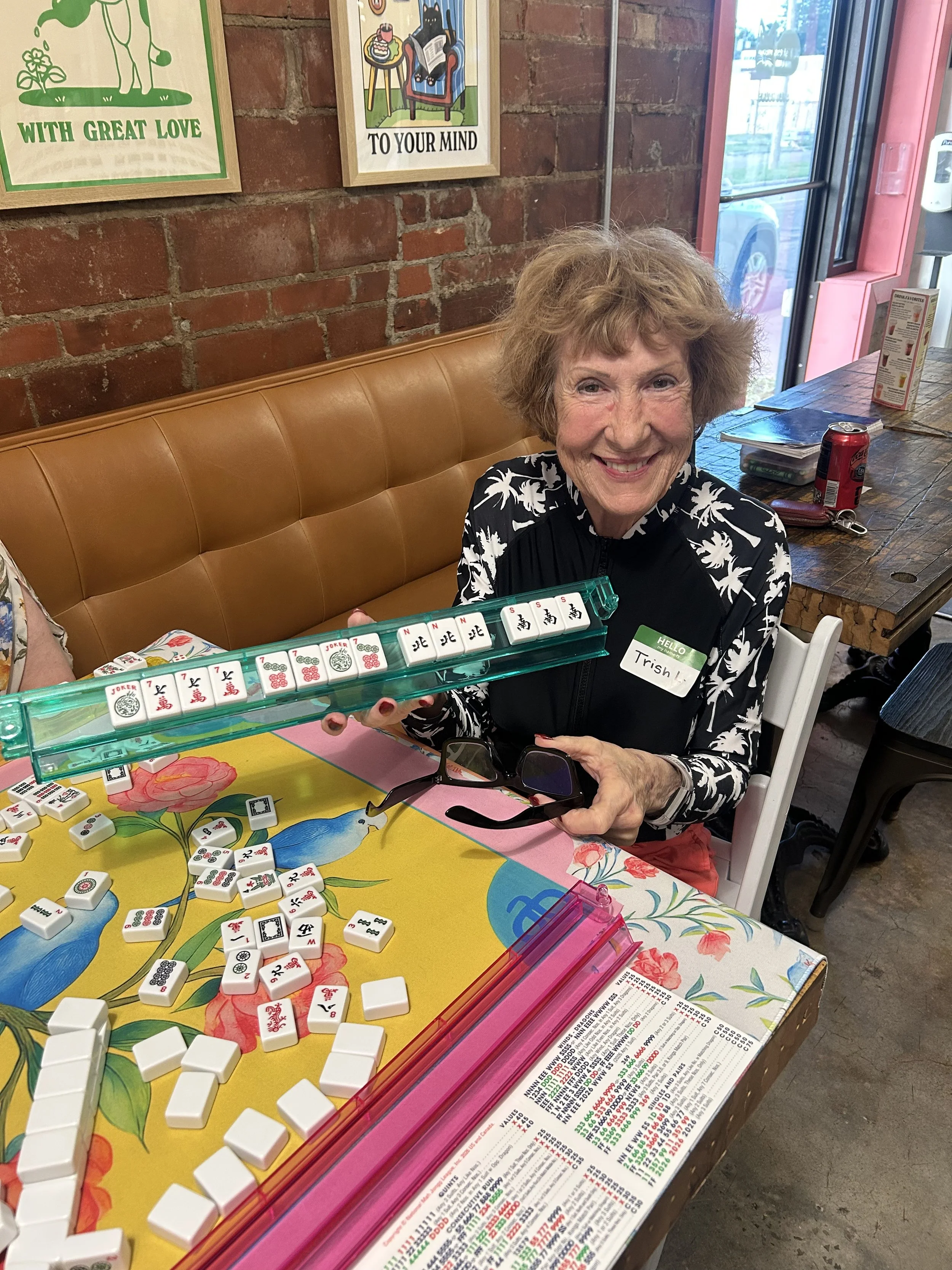 A smiling elderly woman with short, curly hair, wearing a black and white floral jacket, seated at a table playing Mahjong in a casual dining setting. She is holding glasses in one hand and has a name tag reading 'Trish'. The table has Mahjong tiles,