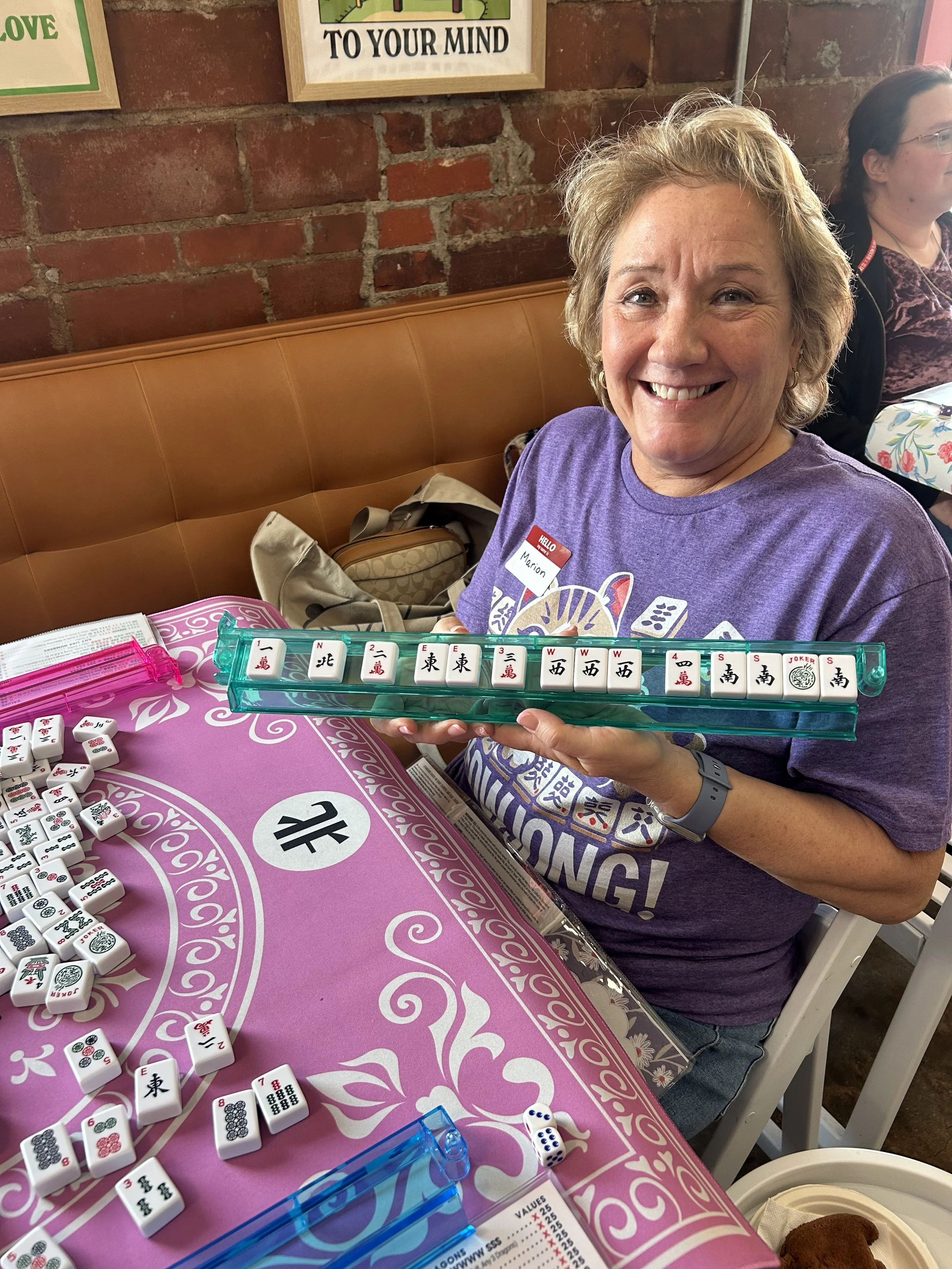 A woman with short blonde hair and a purple T-shirt smiling and holding a row of Mahjong tiles at a game table with pink and white decorative tablecloth. There are additional Mahjong tiles and a die on the table, with a brick wall and framed signs in