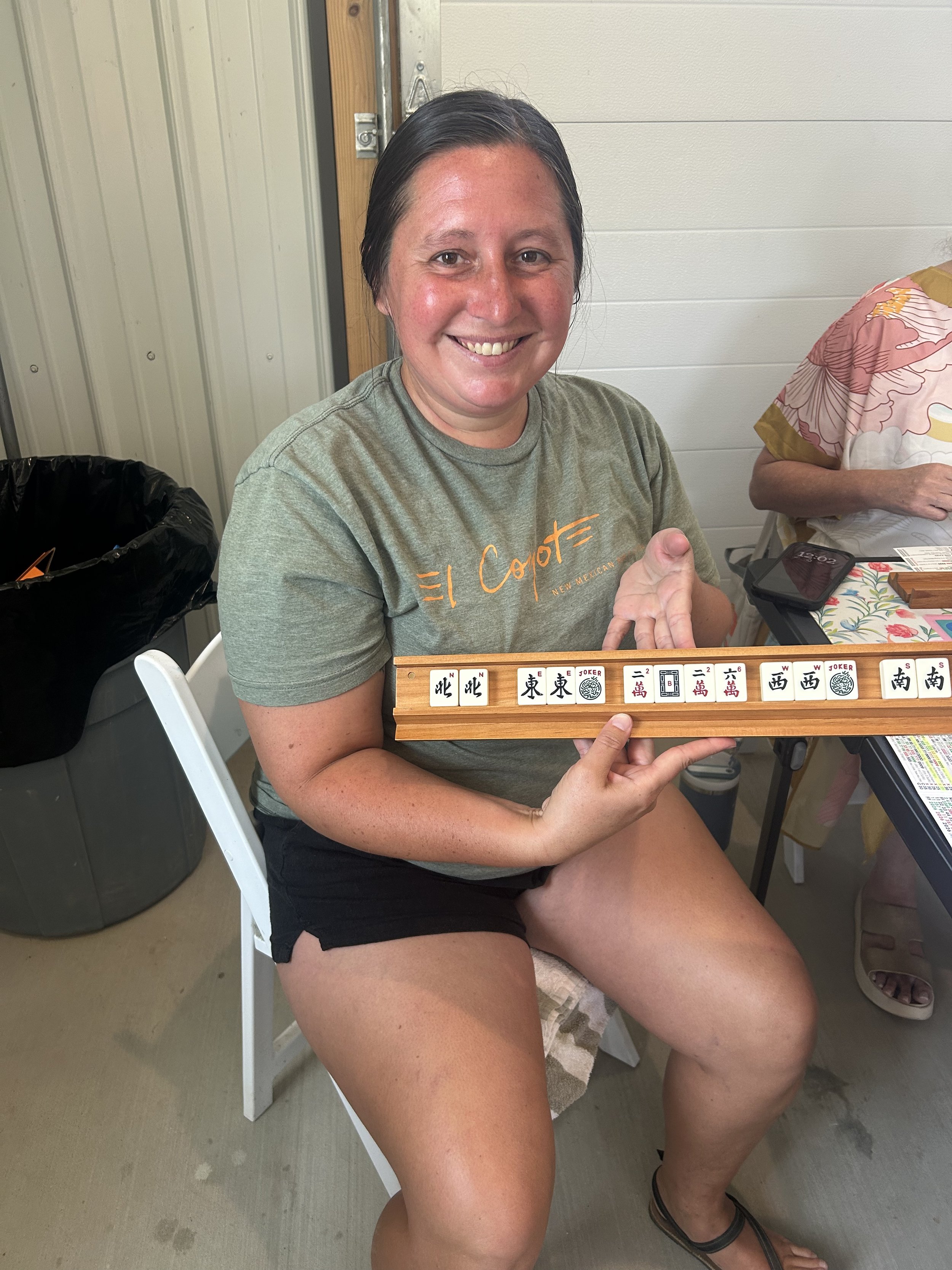 A smiling woman sitting on a white chair, holding a wooden Mahjong tile rack with Mahjong tiles, in a room with a trash can and a table with miscellaneous items, wearing a green T-shirt and black shorts.