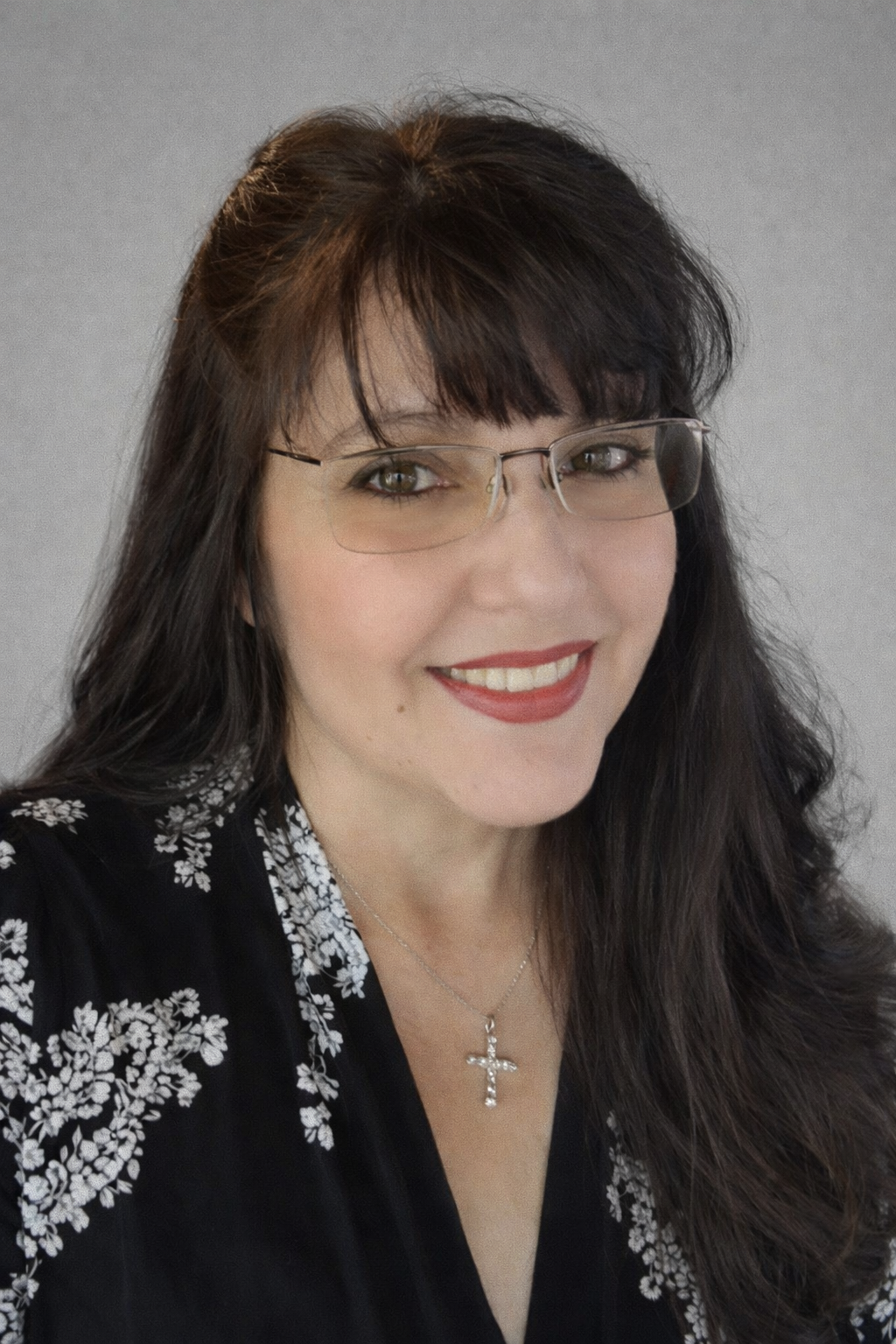 A woman with long dark hair wearing glasses, red lipstick, and a black top with white floral embroidery, smiling, with a gray background.
