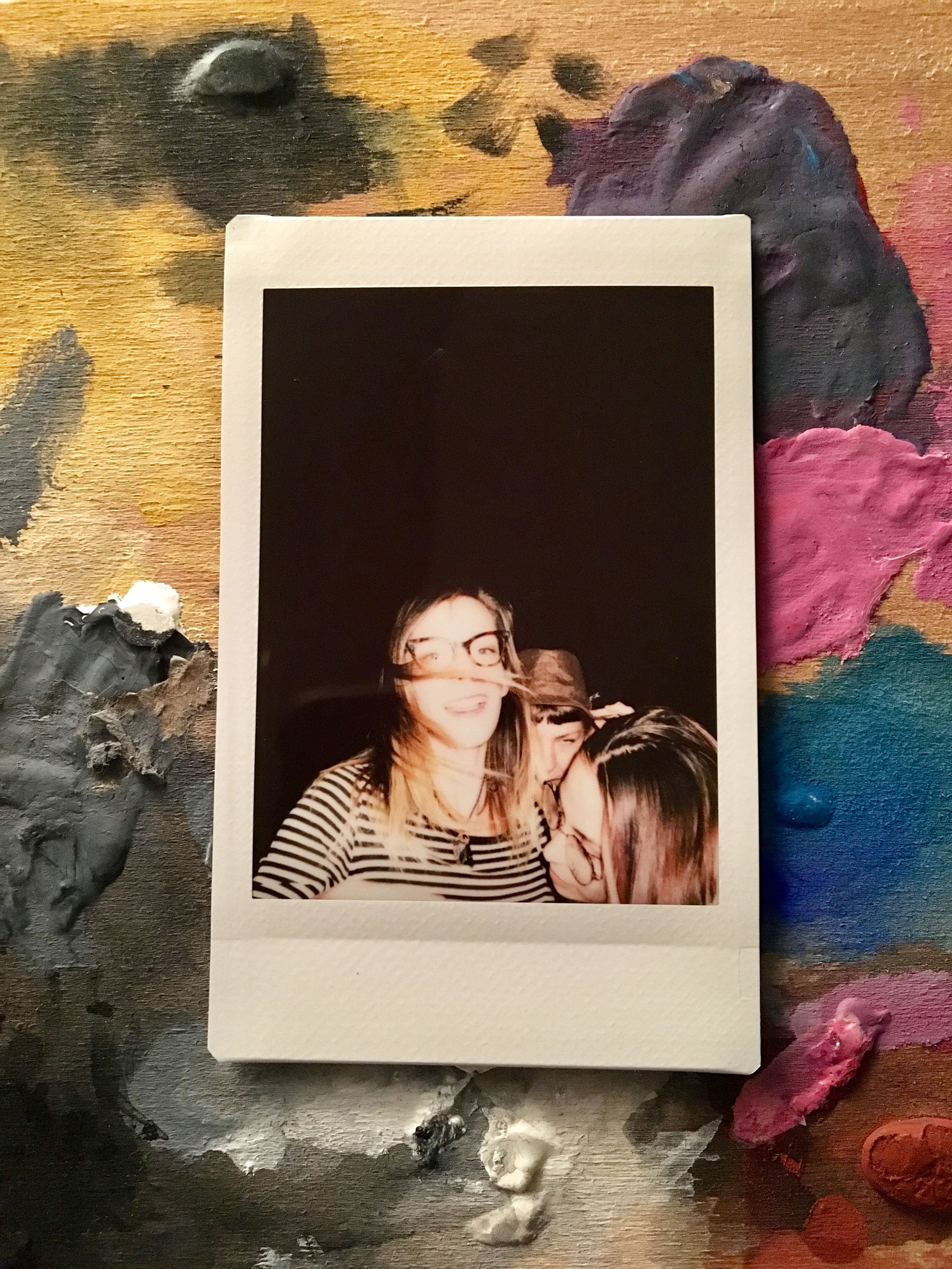 A polaroid photo of two women, one with glasses and a striped shirt, smiling and leaning toward the other, with pink, purple, blue, black, and orange paint on a wooden surface in the background.