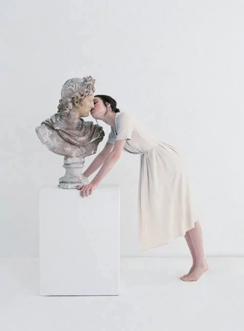 A young girl in a cream dress leans forward, touching and whispering to a classical marble bust of a man, in a minimalist white space.
