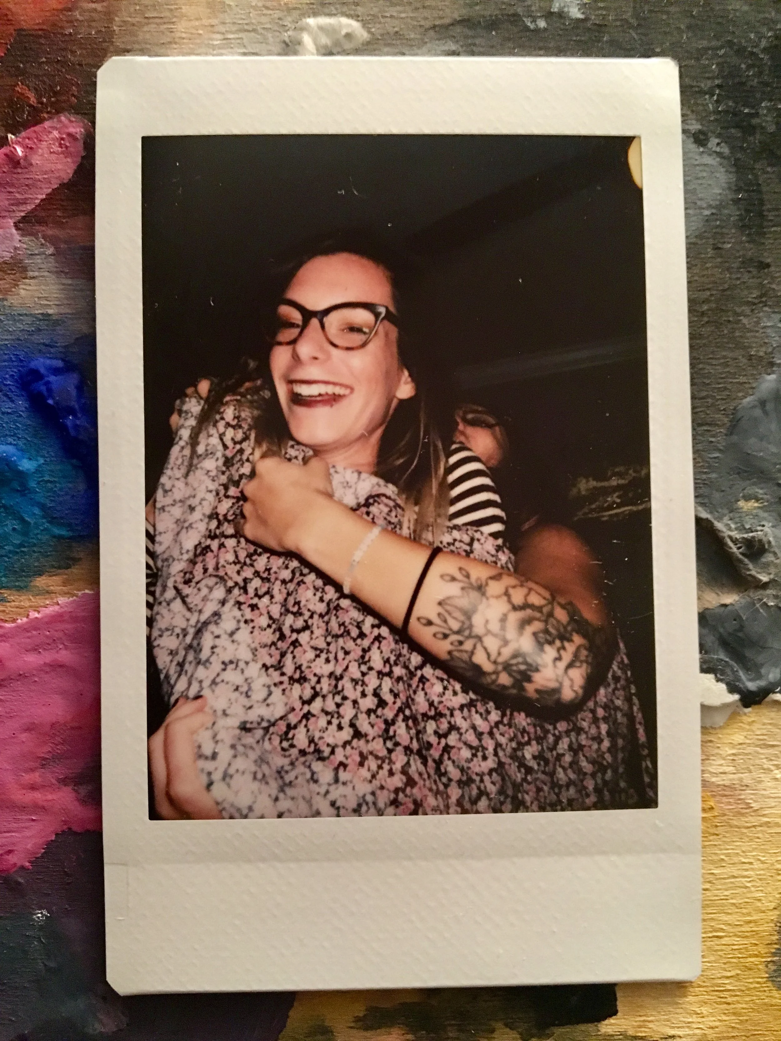 A smiling woman wearing glasses and a patterned shirt hugging someone with a tattooed arm.