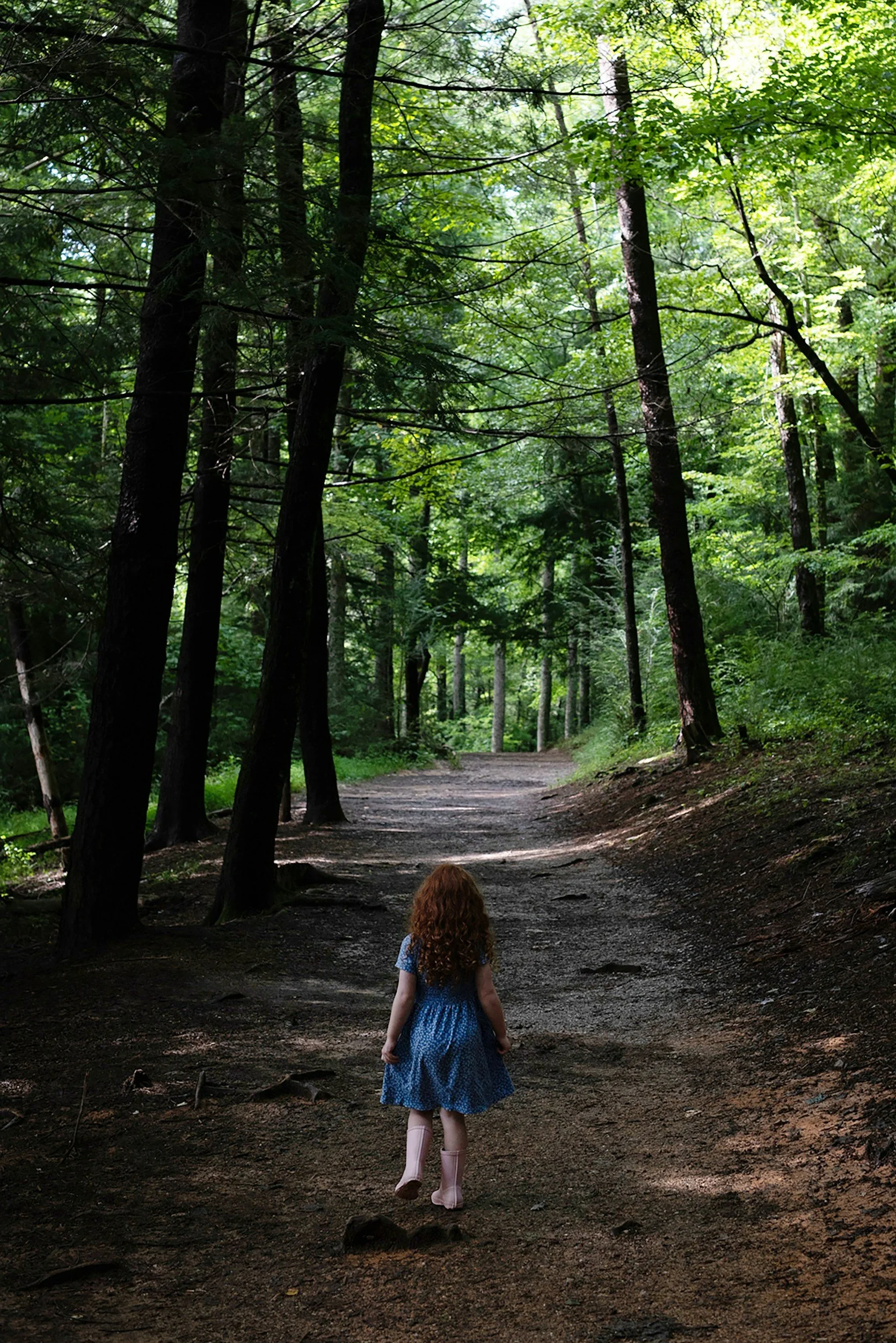 A young girl with curly red hair, wearing a blue dress and pink rain boots, walking down a dirt forest trail surrounded by tall, green trees.