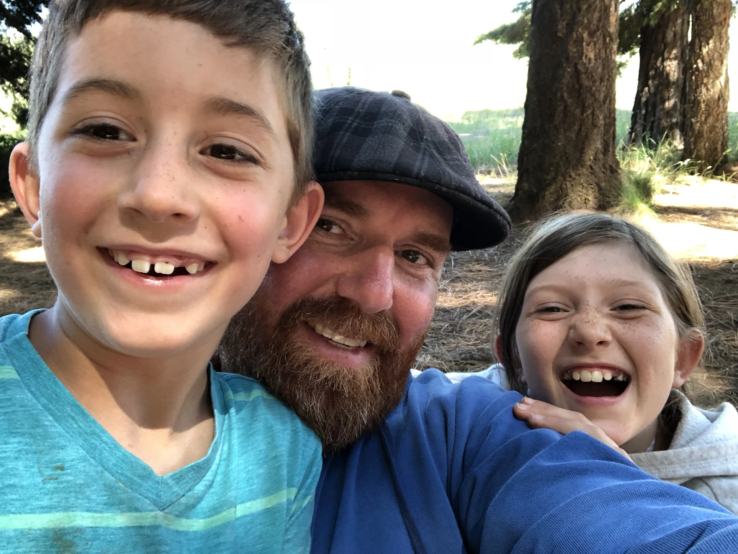 A close-up of a smiling young boy with missing front teeth, an adult man with a beard and a child with long brown hair, all outdoors with trees in the background.