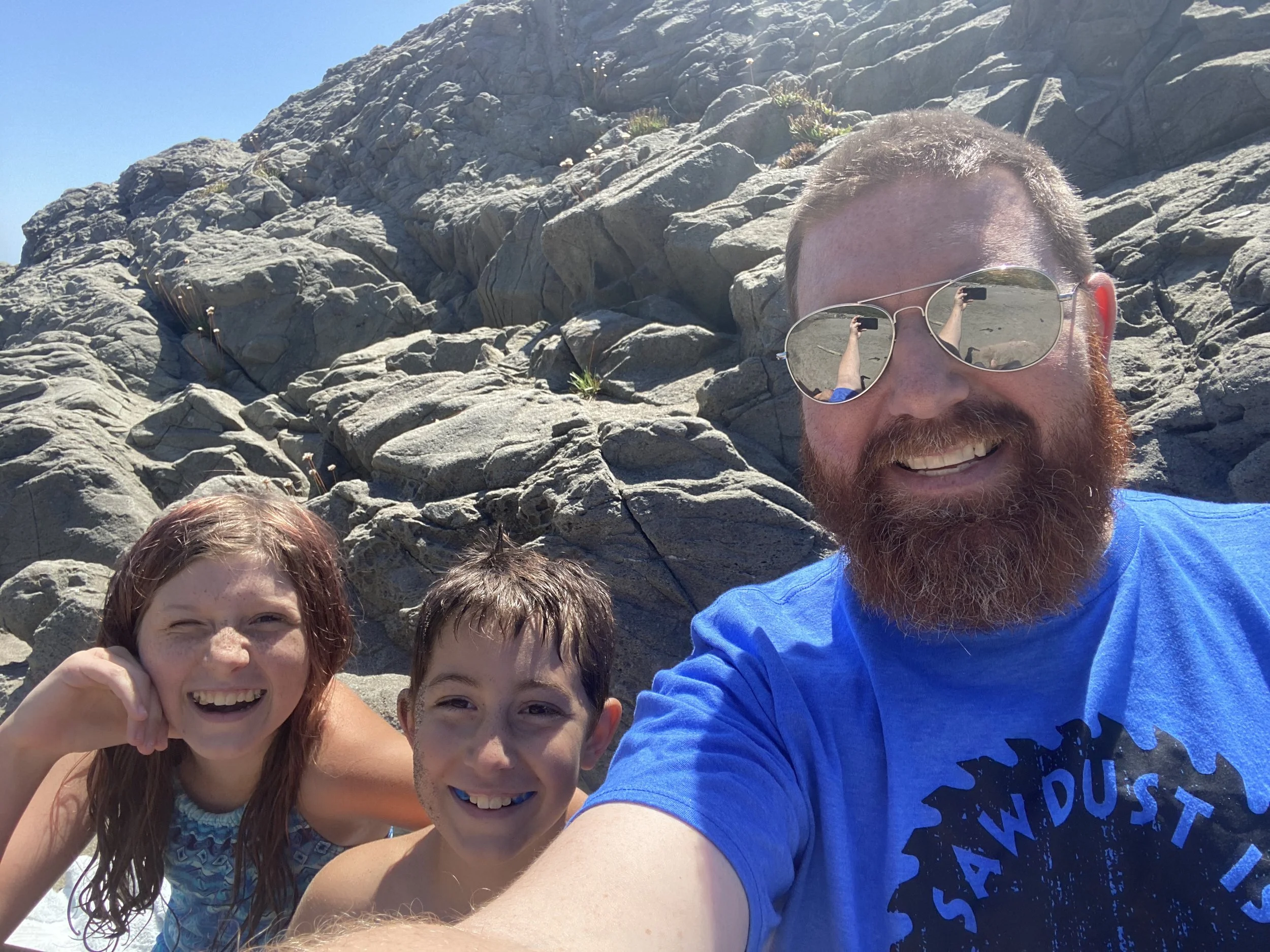 A group of smiling children and a man taking a selfie on a rocky beach under a clear blue sky. The man is wearing sunglasses and a blue t-shirt, and the children are close to him, also smiling.