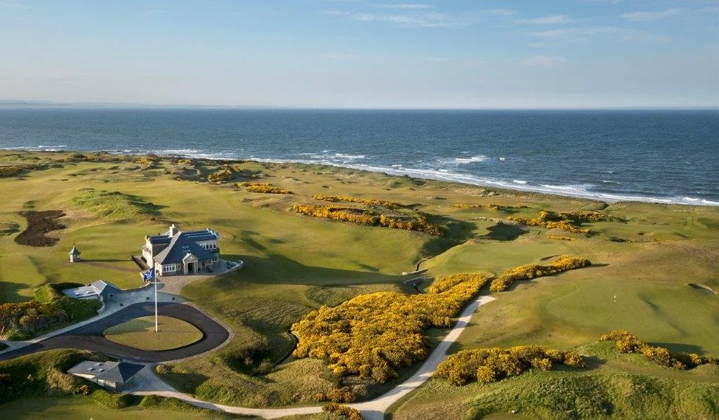 A scenic view of a golf course by the ocean with a large house, a flagpole, and lush green fairways and bushes.