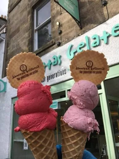 Two ice cream cones with scoops of red and pink ice cream, each topped with a round cookie. The cones are held up in front of an ice cream shop with a sign that reads "Ice Cream & Gelato."