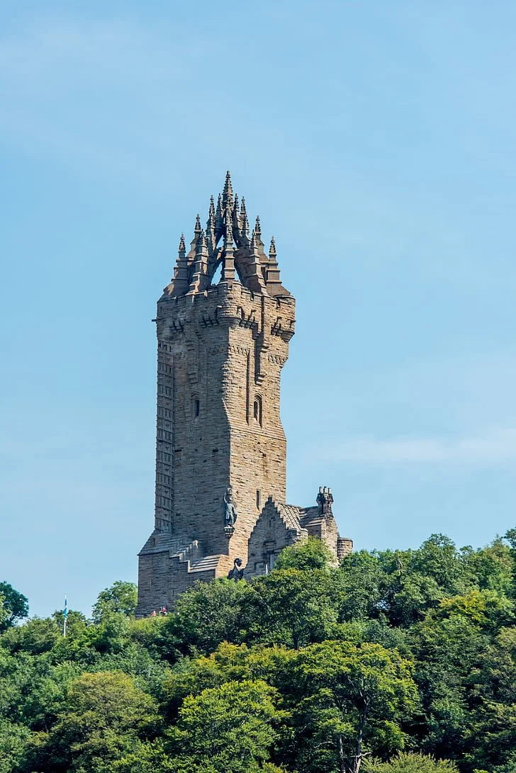 A tall stone castle tower with multiple spires, surrounded by green trees, under a clear blue sky.