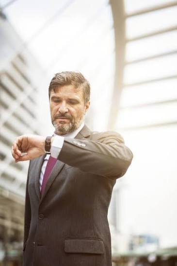 A middle-aged man in a suit looking at his wristwatch outdoors in front of modern architecture with glass and steel elements.
