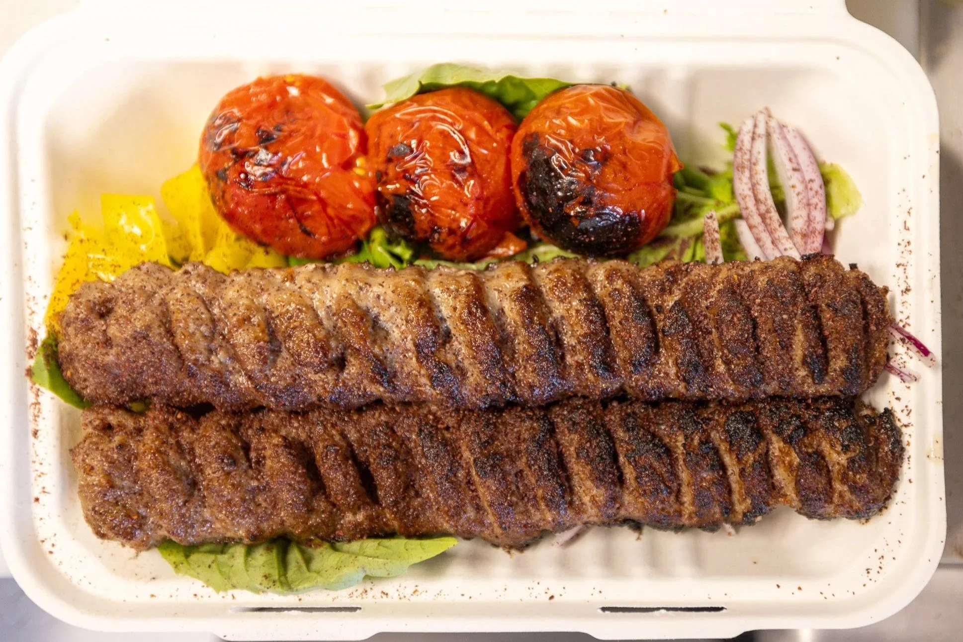 Three grilled kebabs on a bed of lettuce, accompanied by roasted tomatoes, and sliced onions with spices, in a takeout container.