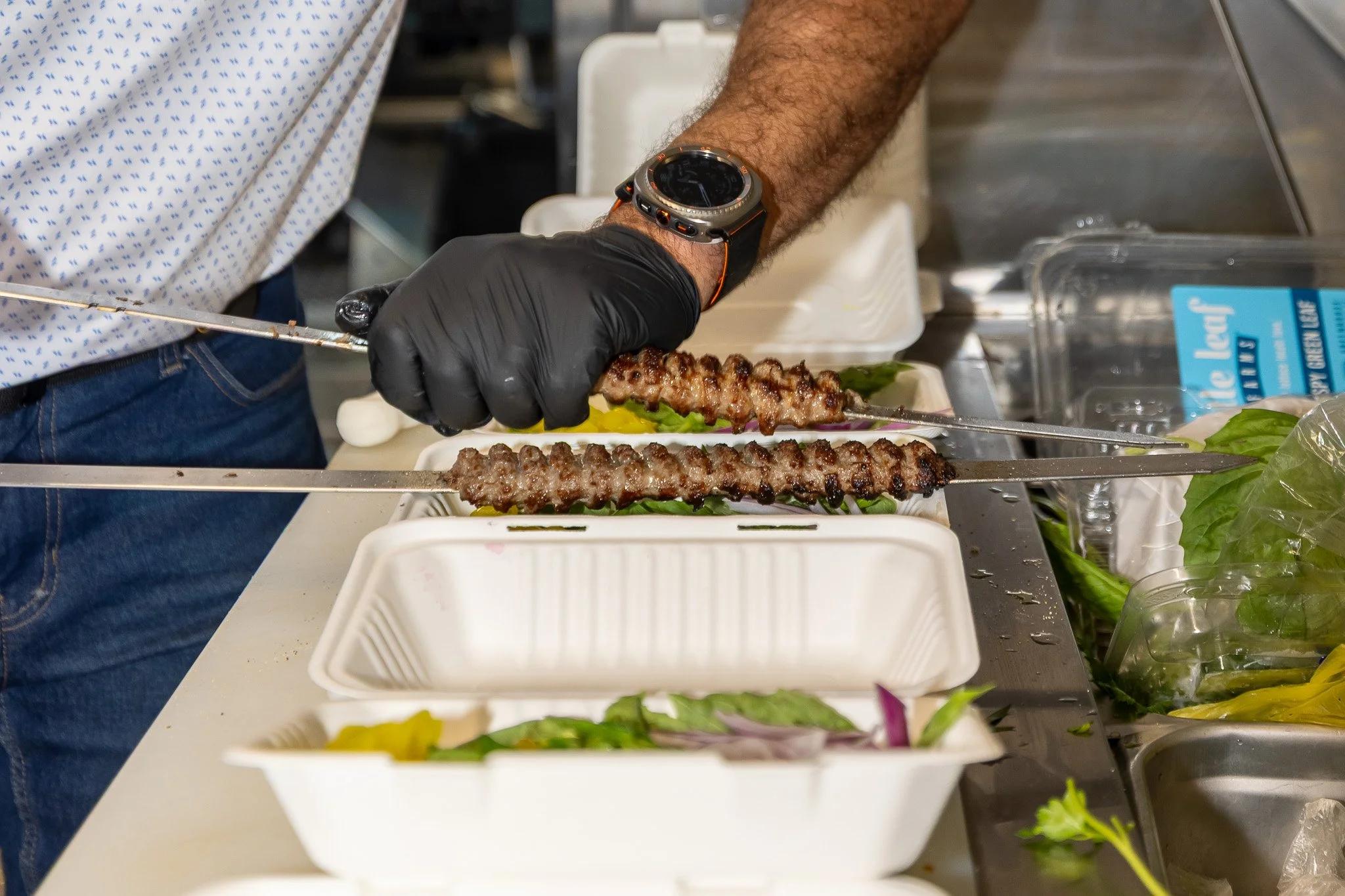 A person with a black glove and an orange strap smartwatch is skewering grilled meat on metal skewers, preparing food in a kitchen or food stall. There are stacked white containers, fresh greens, and a plastic water bottle nearby.