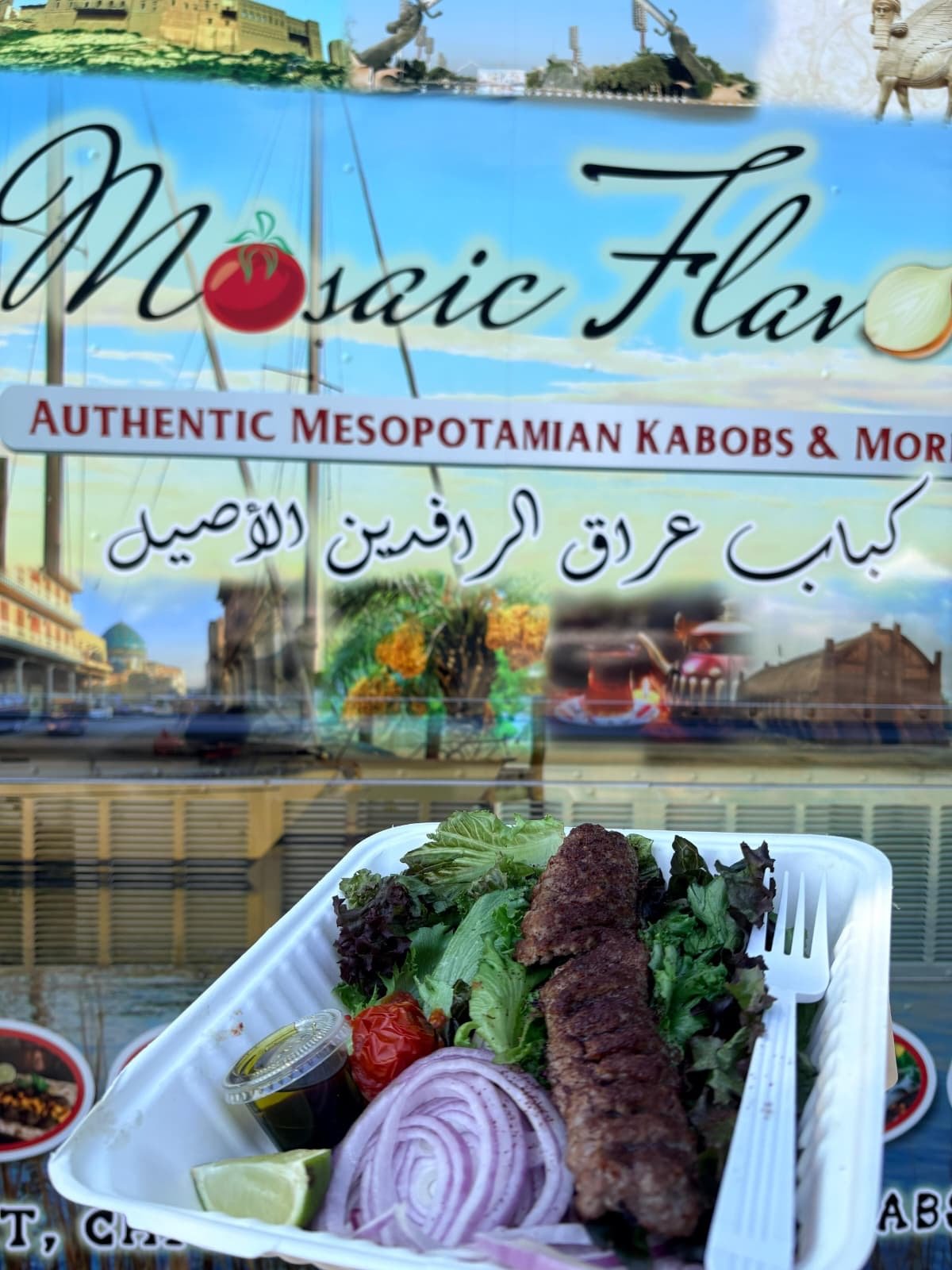 A tray of grilled kebabs with fresh salad, onions, tomato, lemon, and a small container of sauce in front of a sign for a restaurant called Mosaic Flavors, serving authentic Mesopotamian kebabs and more.
