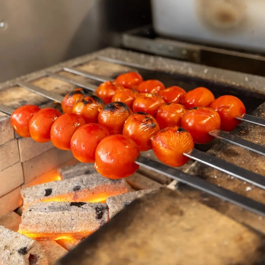 Charred tomatoes on metal skewers placed over a brick and fire grill.