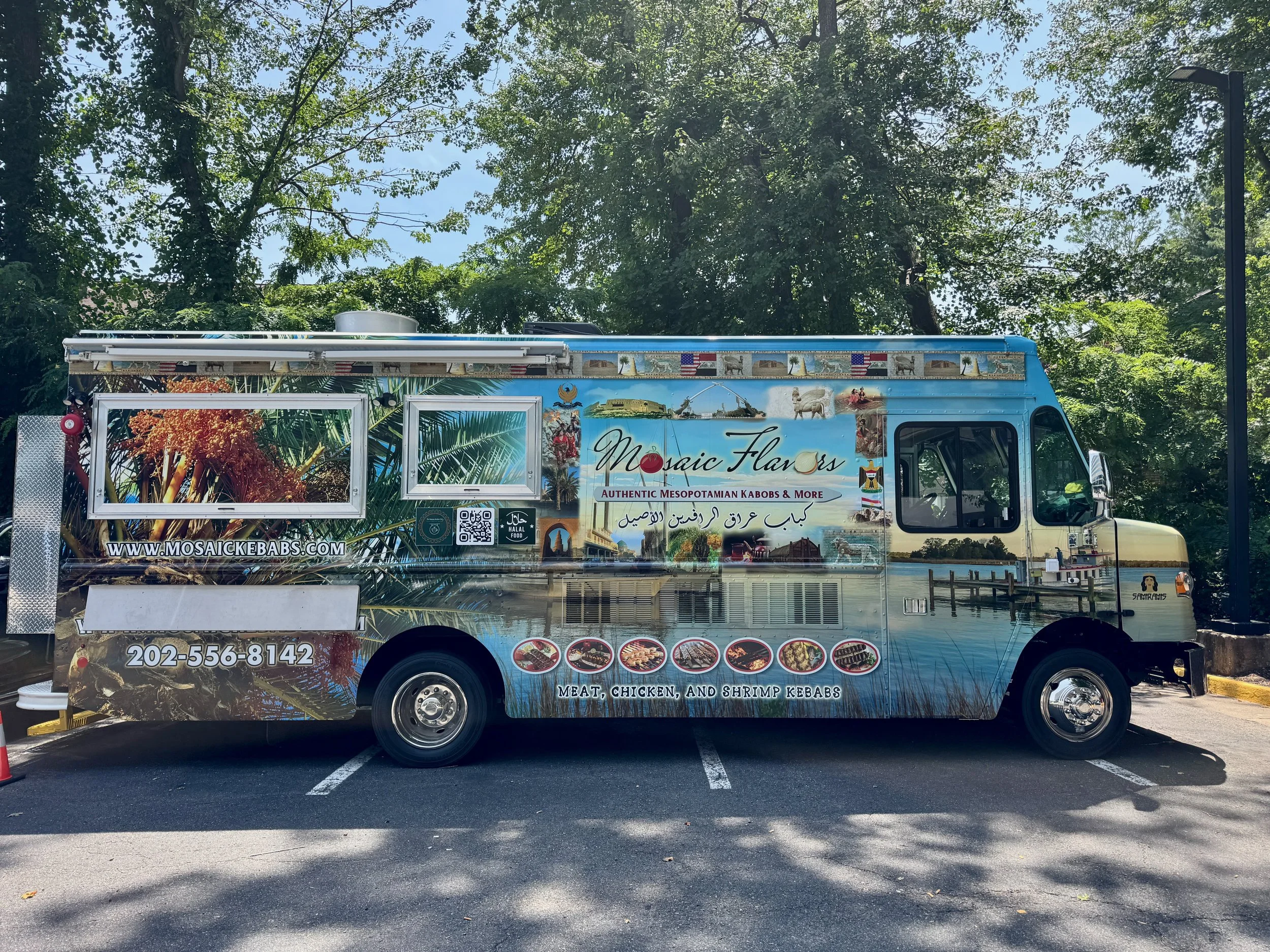 Side view of a food truck selling Middle Eastern kebabs, with colorful signage displaying images of meat, chicken, and shrimp kebabs, along with the restaurant's website and phone number, parked in an outdoor lot under tall trees.