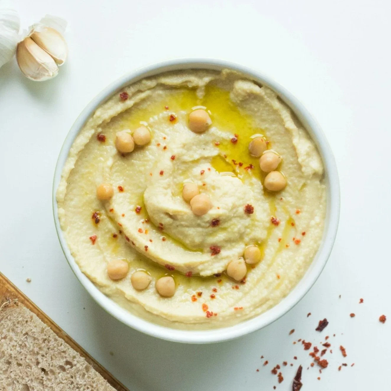 A bowl of hummus garnished with whole chickpeas, red pepper flakes, and olive oil, on a white surface with garlic cloves nearby and some crumbs and herbs.
