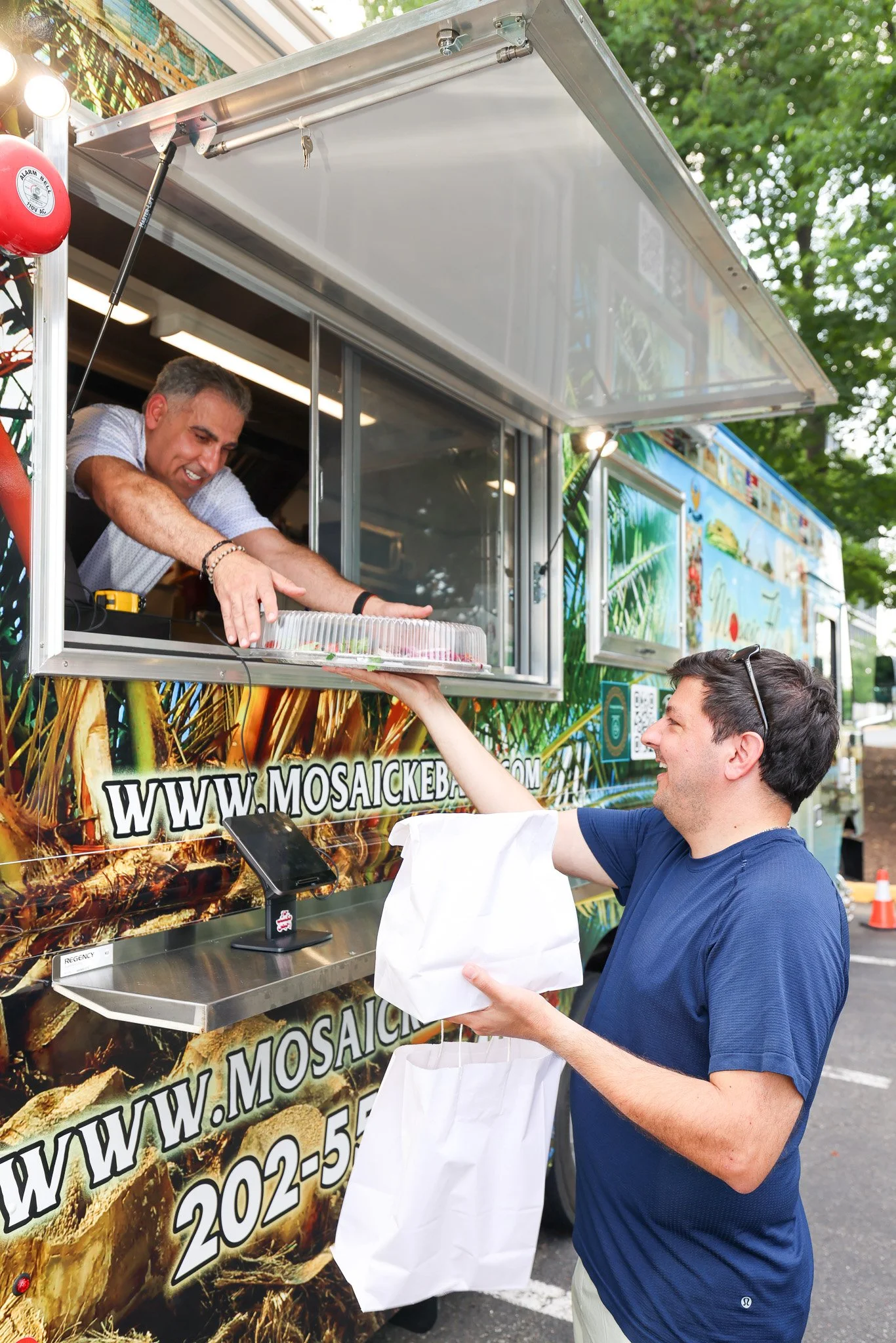 A man in a blue shirt receives a package from a man inside a food truck, which has tropical-themed decorations and a website URL on it.