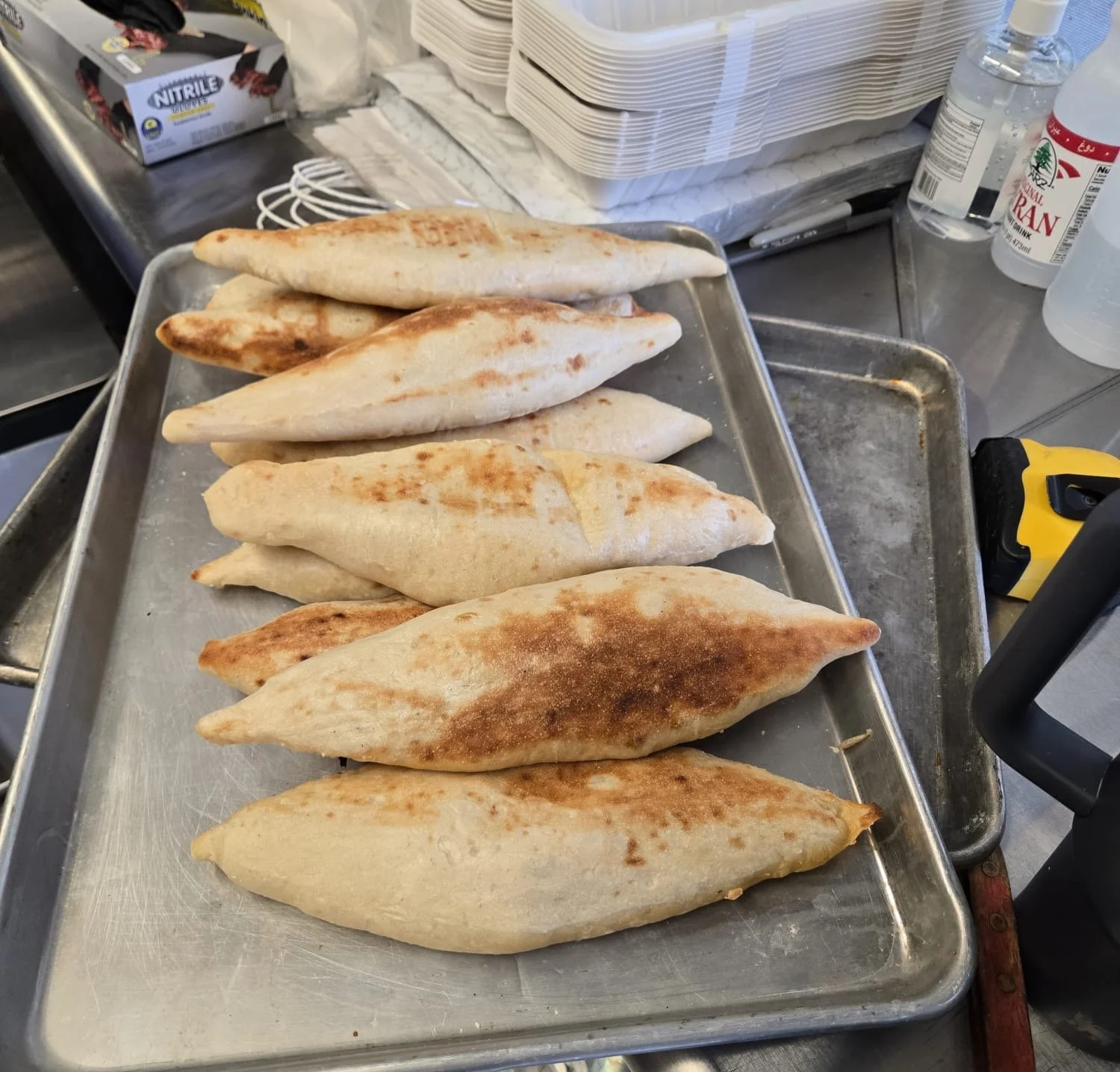 A metal tray with seven freshly baked, golden-brown, Samoon (traditional Iraqi bread)  on a stainless steel countertop in a food truck kitchen.
