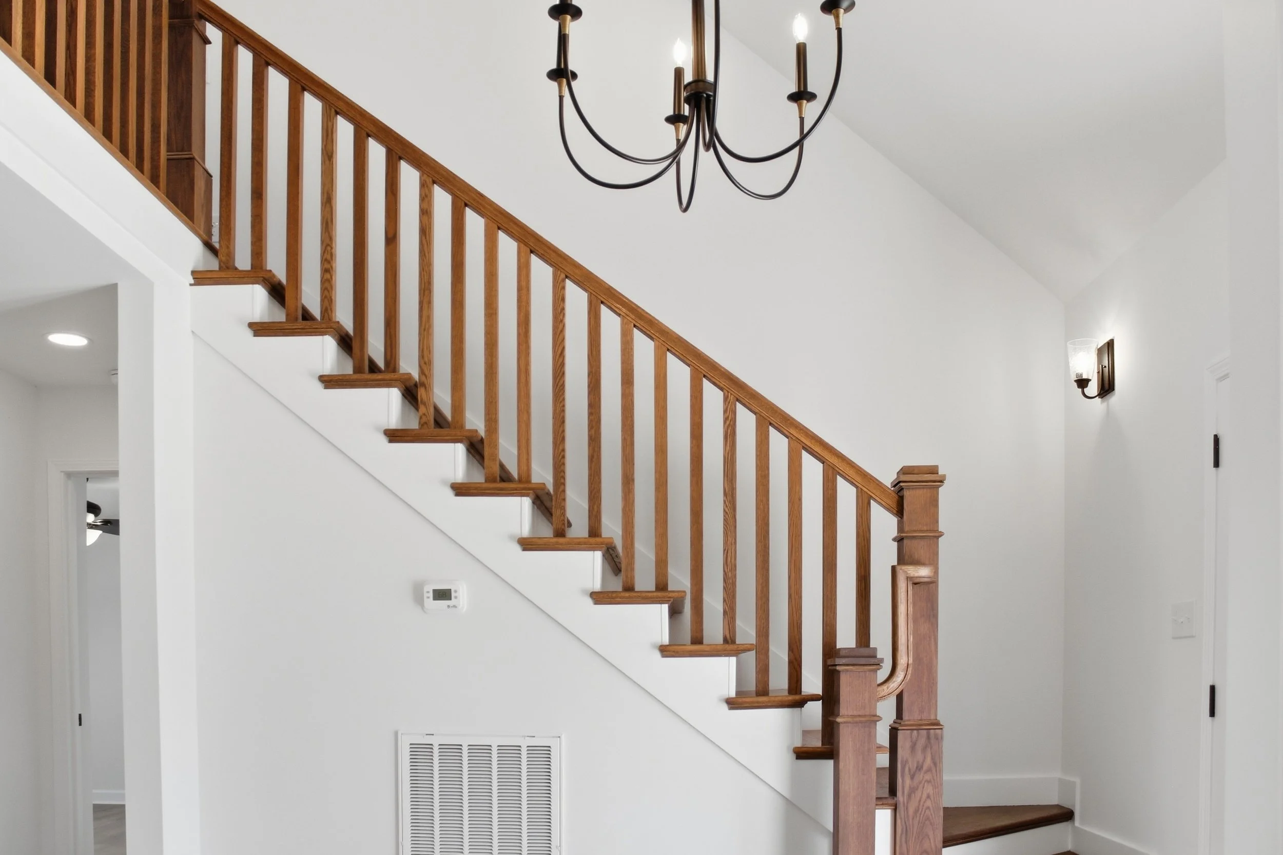 Interior view of a staircase with wooden handrails and white risers, a chandelier hanging from the ceiling, and wall-mounted light fixtures on a white wall.