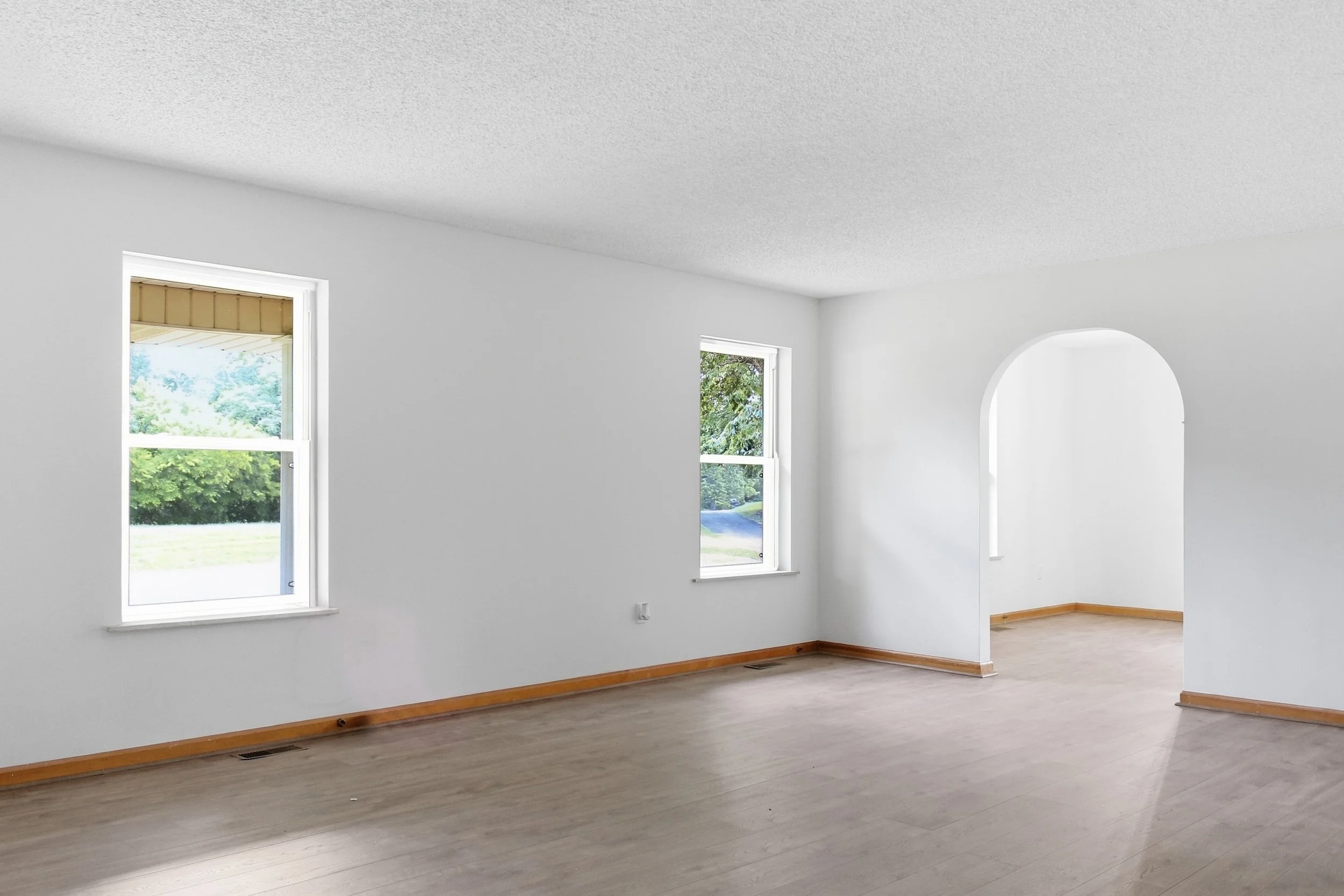 Empty room with white walls, two windows showing greenery outside, wooden floor, and a small archway.