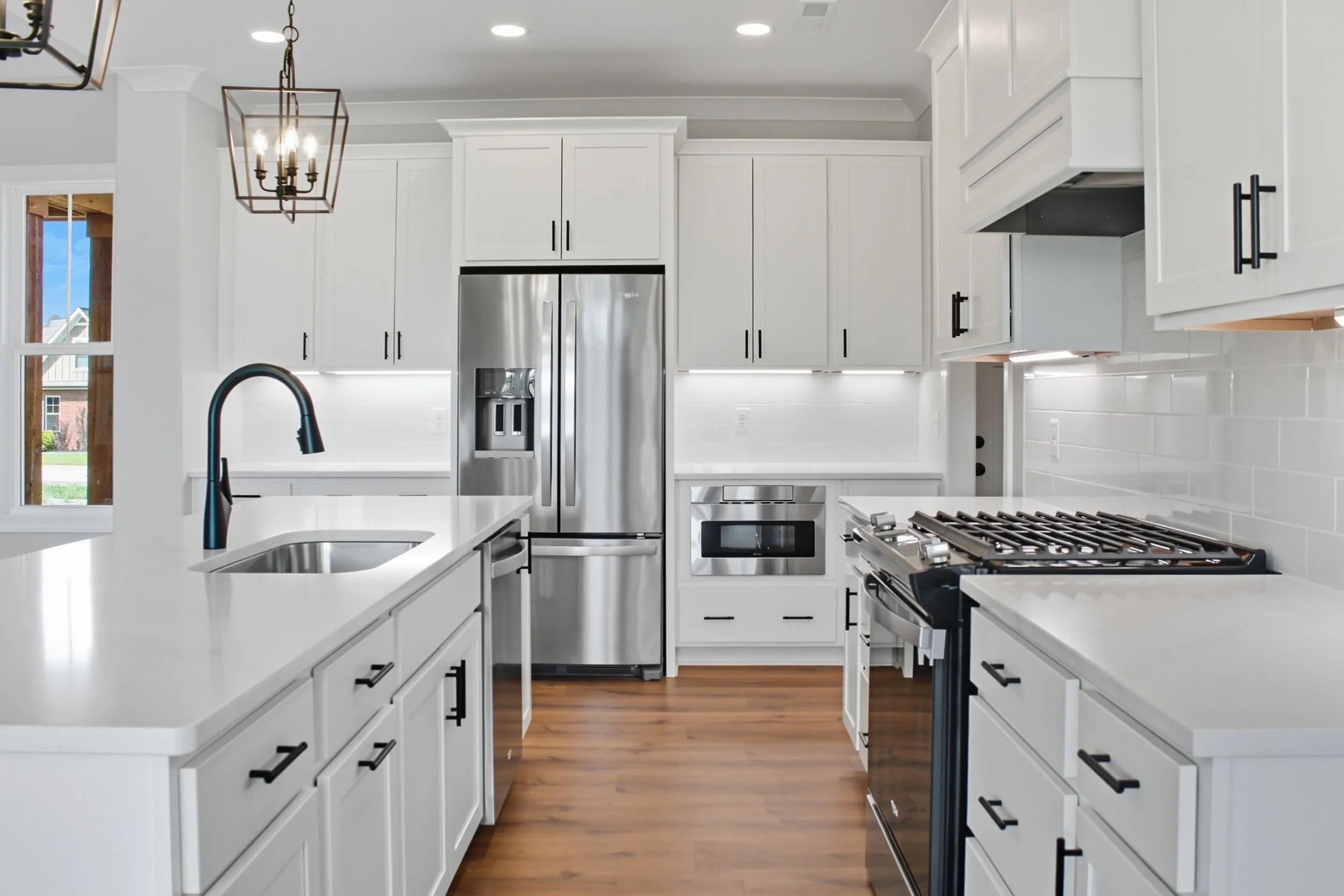 Modern white kitchen with black cabinet handles, stainless steel refrigerator, gas stove, and black faucet, hardwood floors, and a window showing a suburban neighborhood.
