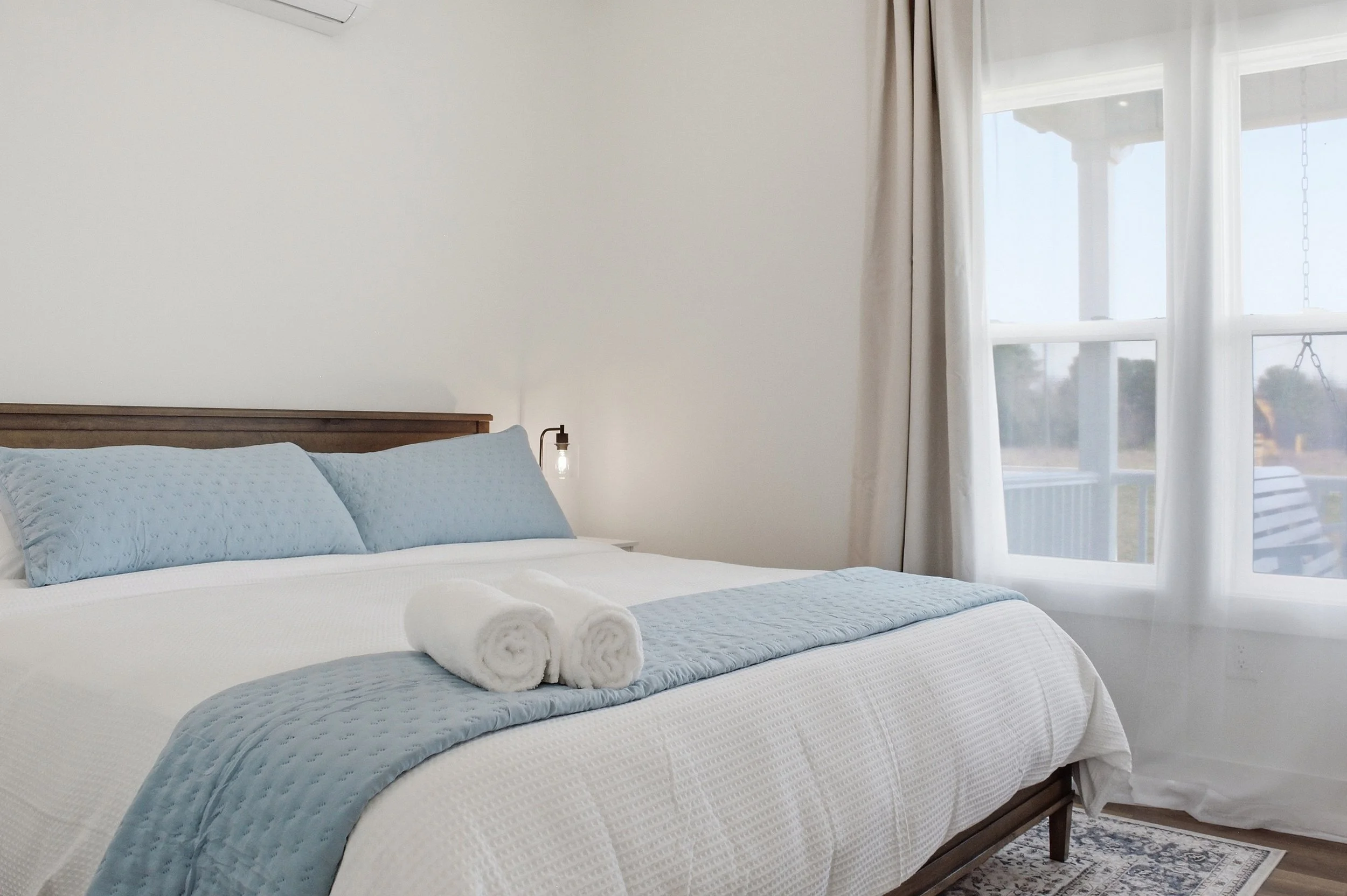 A neatly made bed with white and light blue bedding, two rolled white towels on top, in a bright bedroom with white walls, a large window with white curtains, and a side lamp.