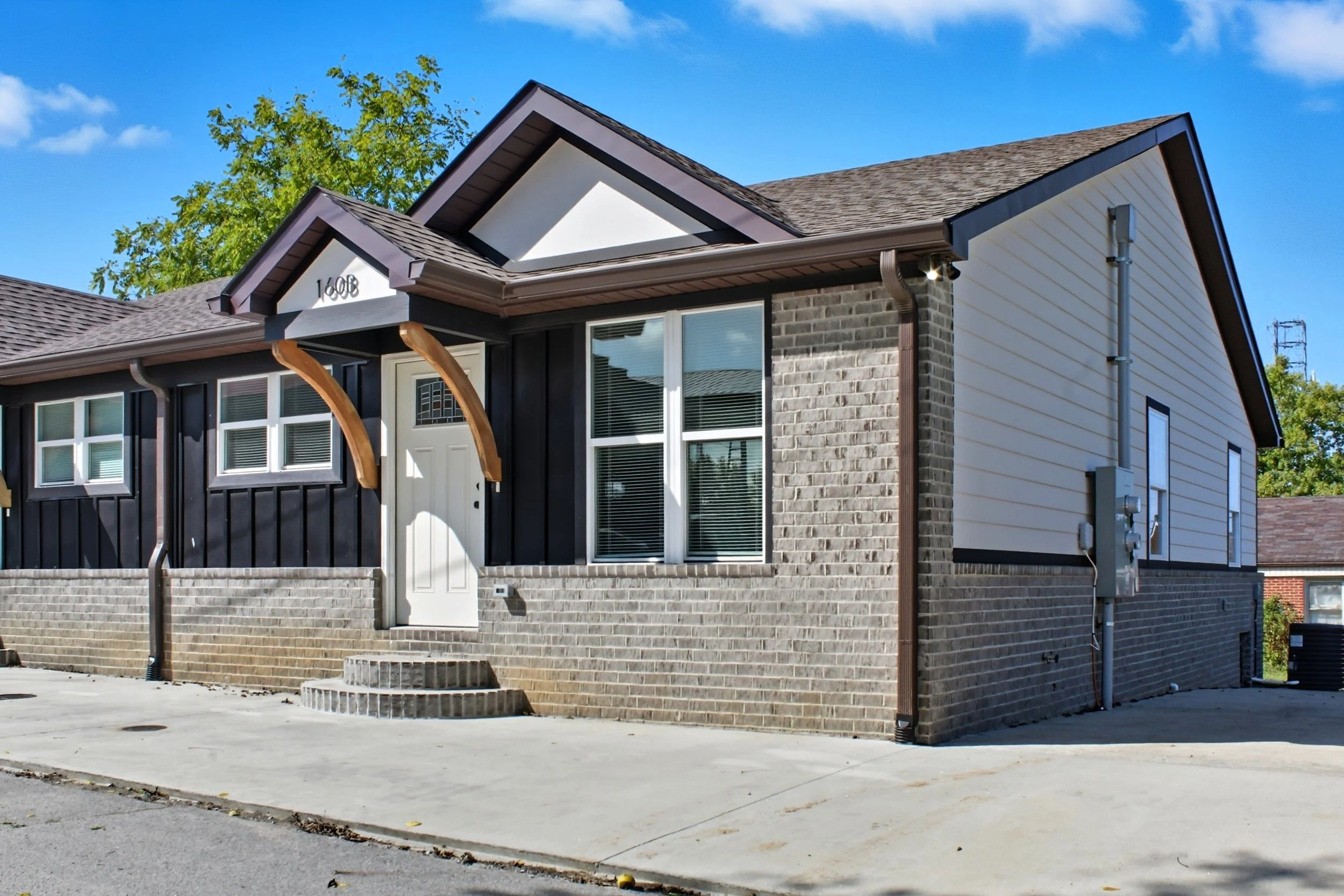 A modern two-story house with a slanted roof, white siding, dark wood accents, and large windows. There is a small set of stairs leading to the front door and a concrete driveway in front. A tree with green leaves is visible behind the house, and the sky is blue with some clouds.