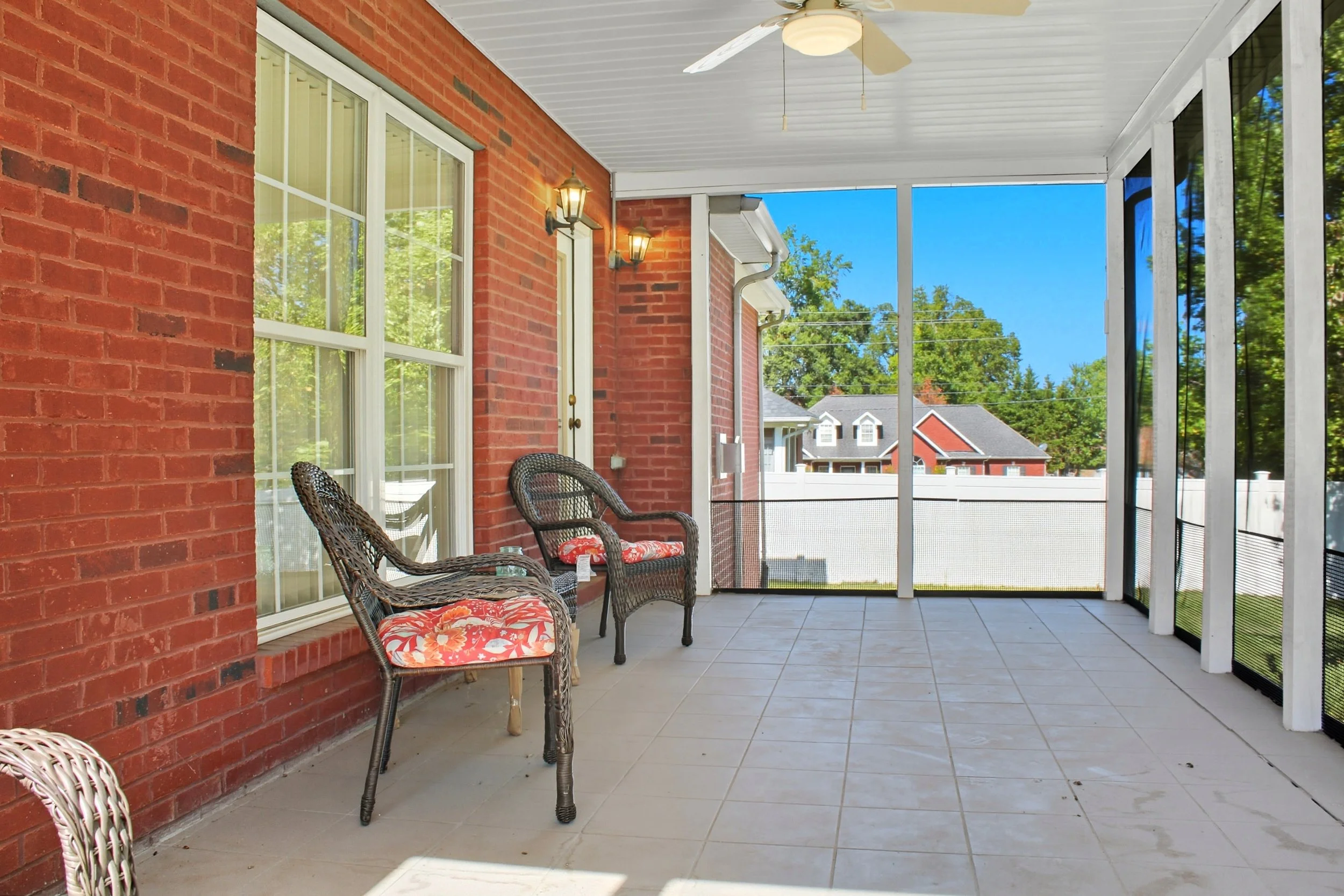 Back porch area with brick wall, large sliding glass door, ceiling fan, and outdoor seating with four chairs featuring orange floral cushions, overlooking a neighborhood with trees and houses under a blue sky.