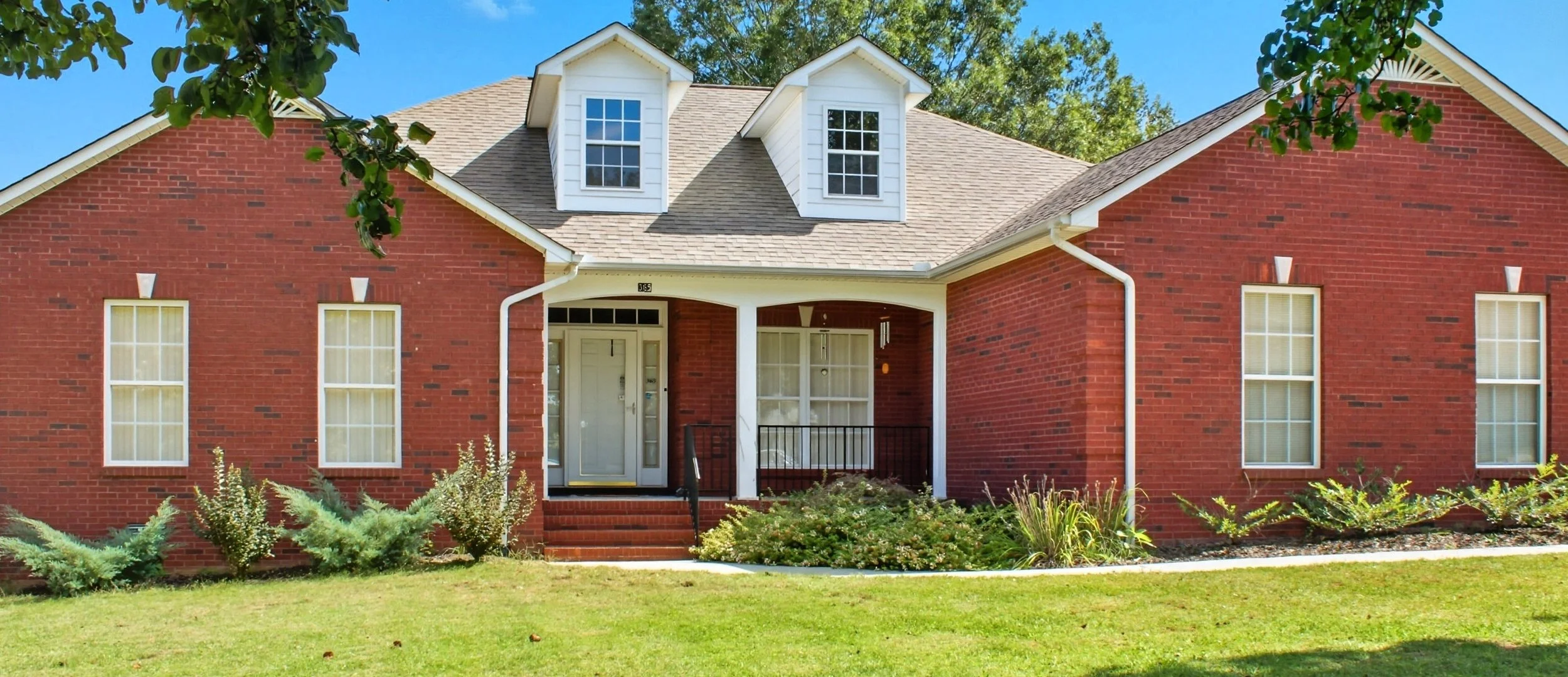 Red brick house with white trim, two small dormer windows on the roof, a front porch with steps, a white front door, and multiple windows with yellow curtains. Green grass and bushes in front, under a blue sky.