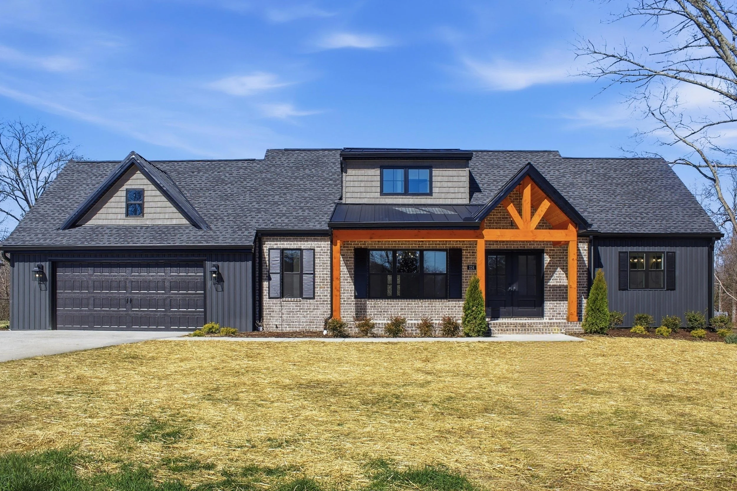 Front view of a modern house with brick and dark siding, covered porch with wooden beams, a large garage, and a lawn with sparse grass and small shrubs.