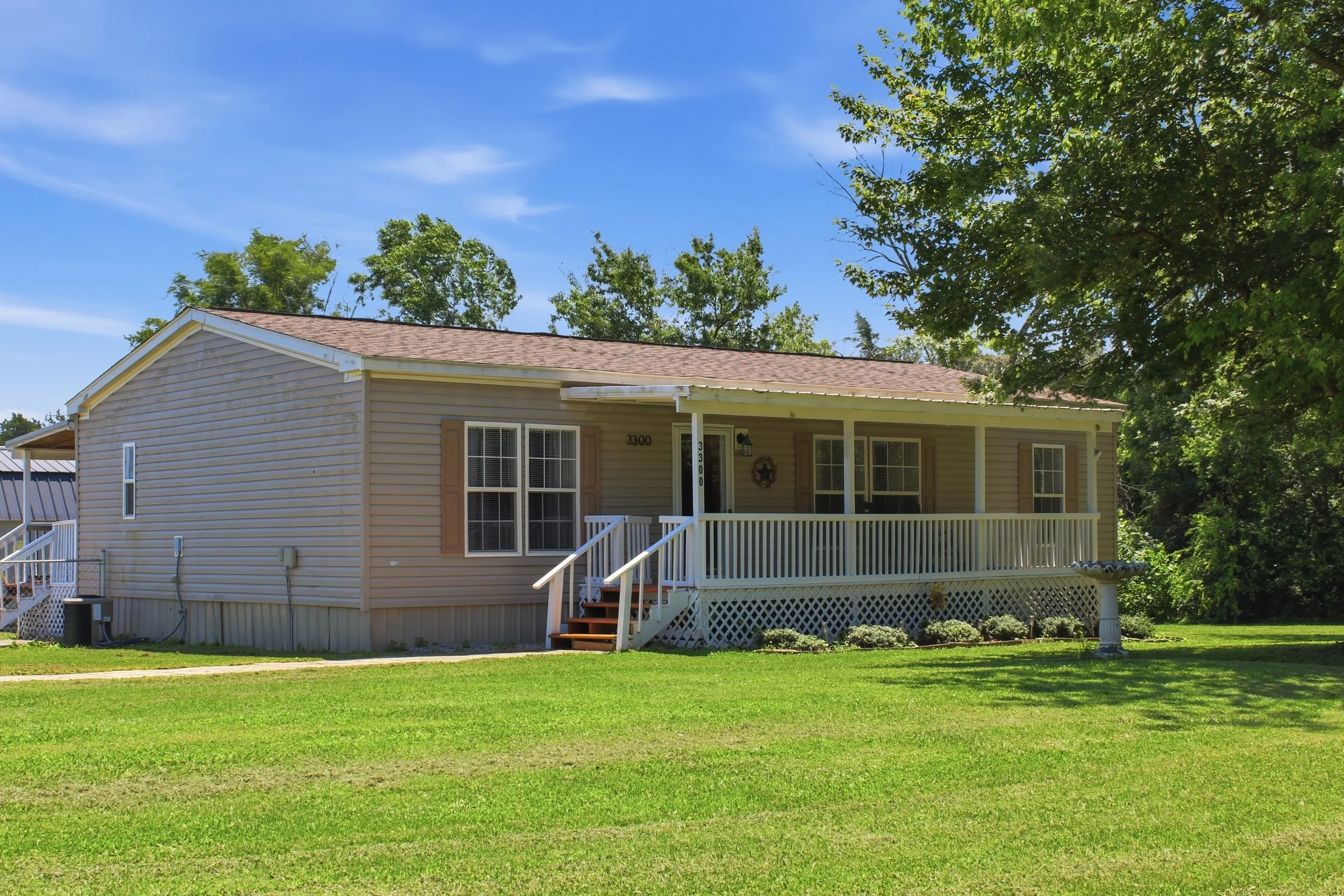 A beige house with a front porch, white railings, and brown steps, set on a green lawn with trees and a blue sky in the background.