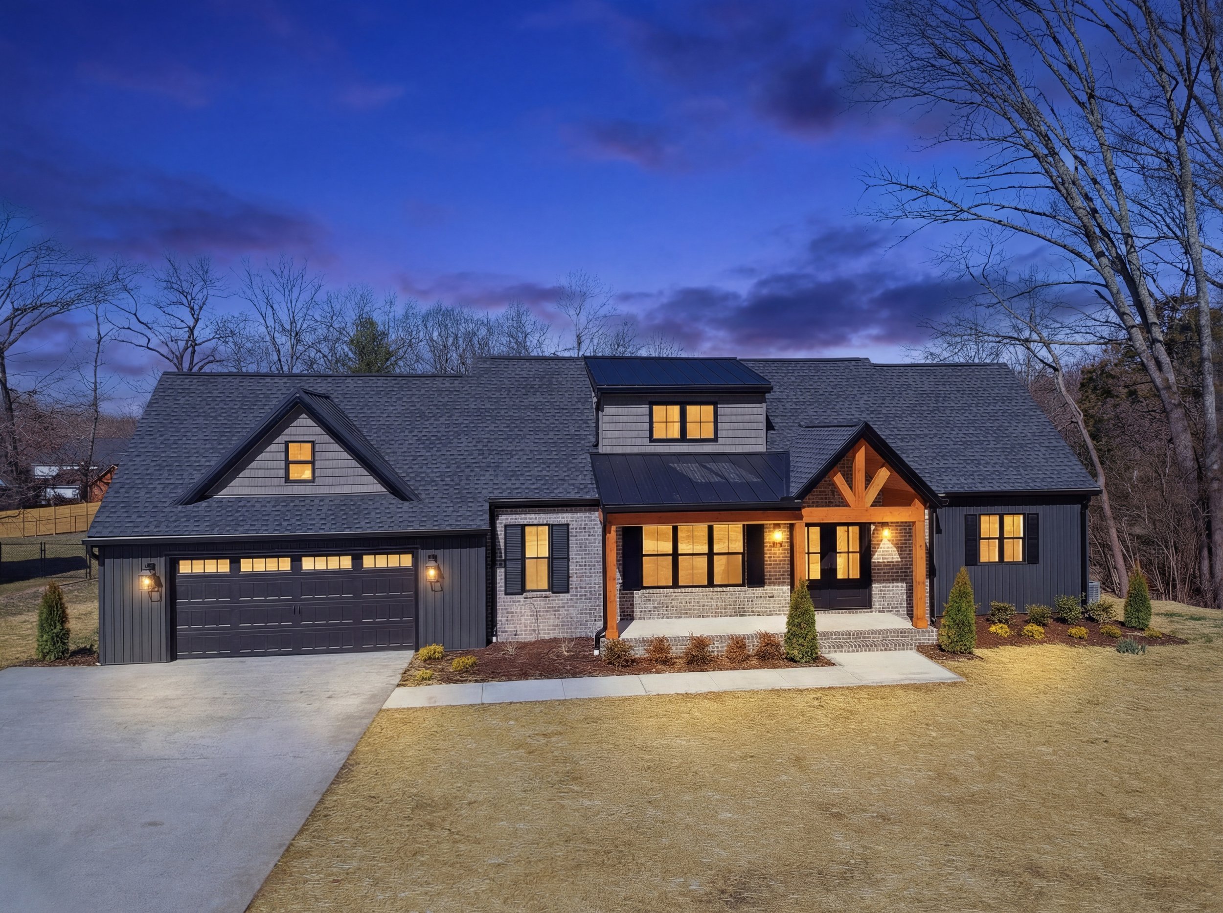 Modern house with large windows and illuminated interior lights, a garage door, front porch with wooden support beams, well-manicured lawn and shrubs, leafless trees in background, evening sky with dark clouds.