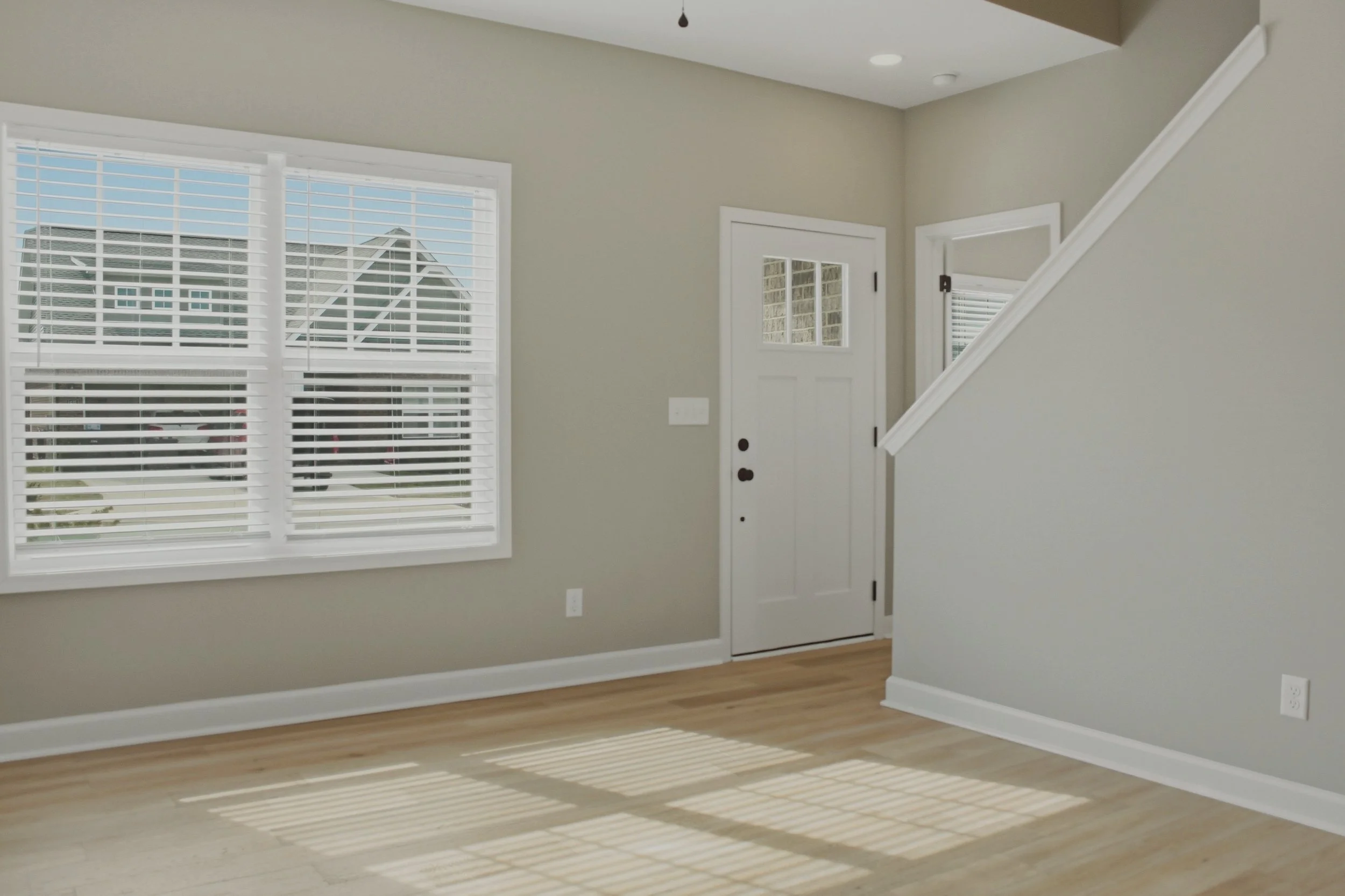 Interior of a house with a window with white blinds, a front door, and a staircase. The floor is light wood, and sunlight creates shadow lines on the floor.